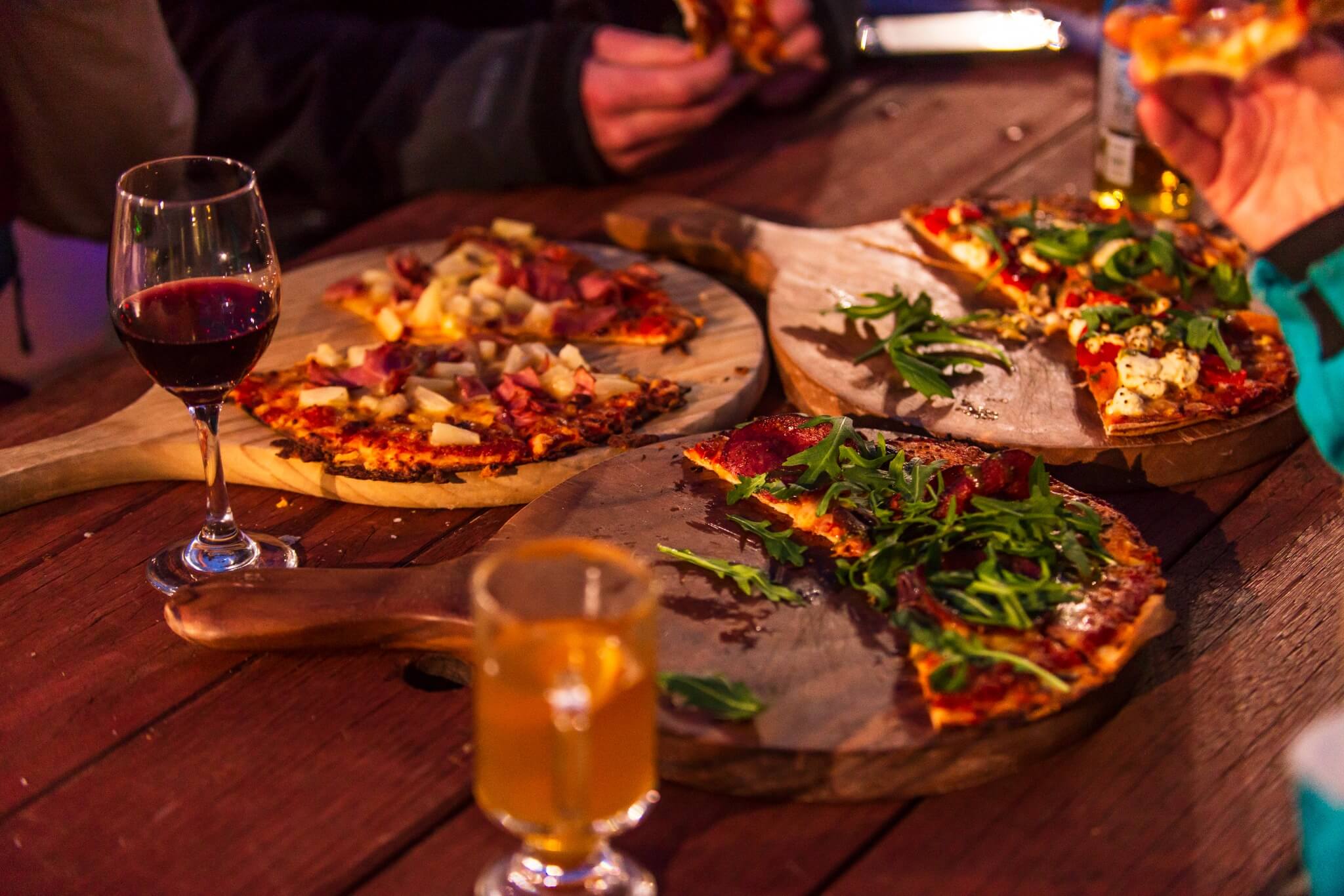 Three wooden boards with sliced pizza topped with greens, beside glasses of red and white drinks.