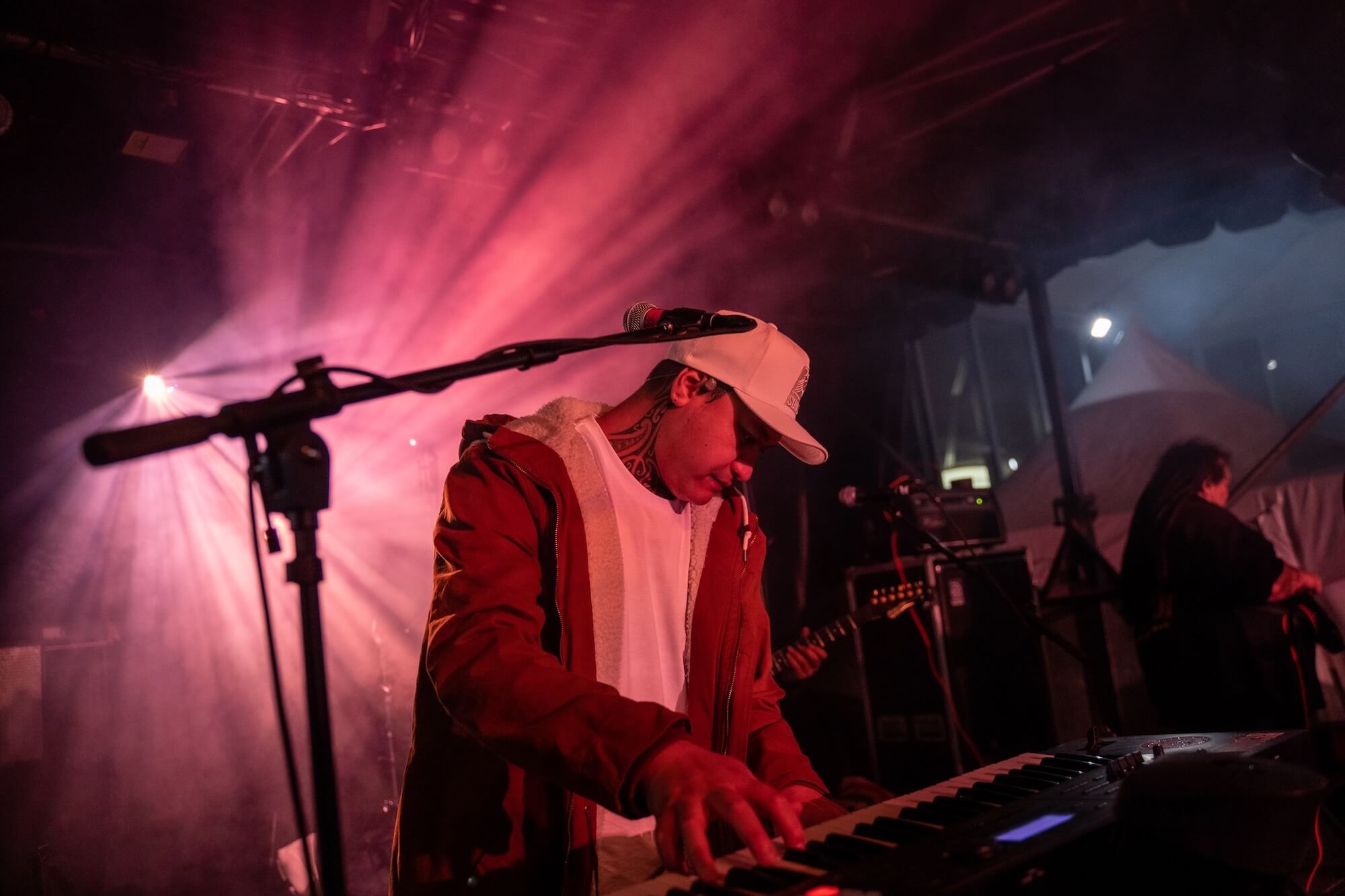 Musician wearing a red jacket and white cap playing keyboard on stage with pink light beams.