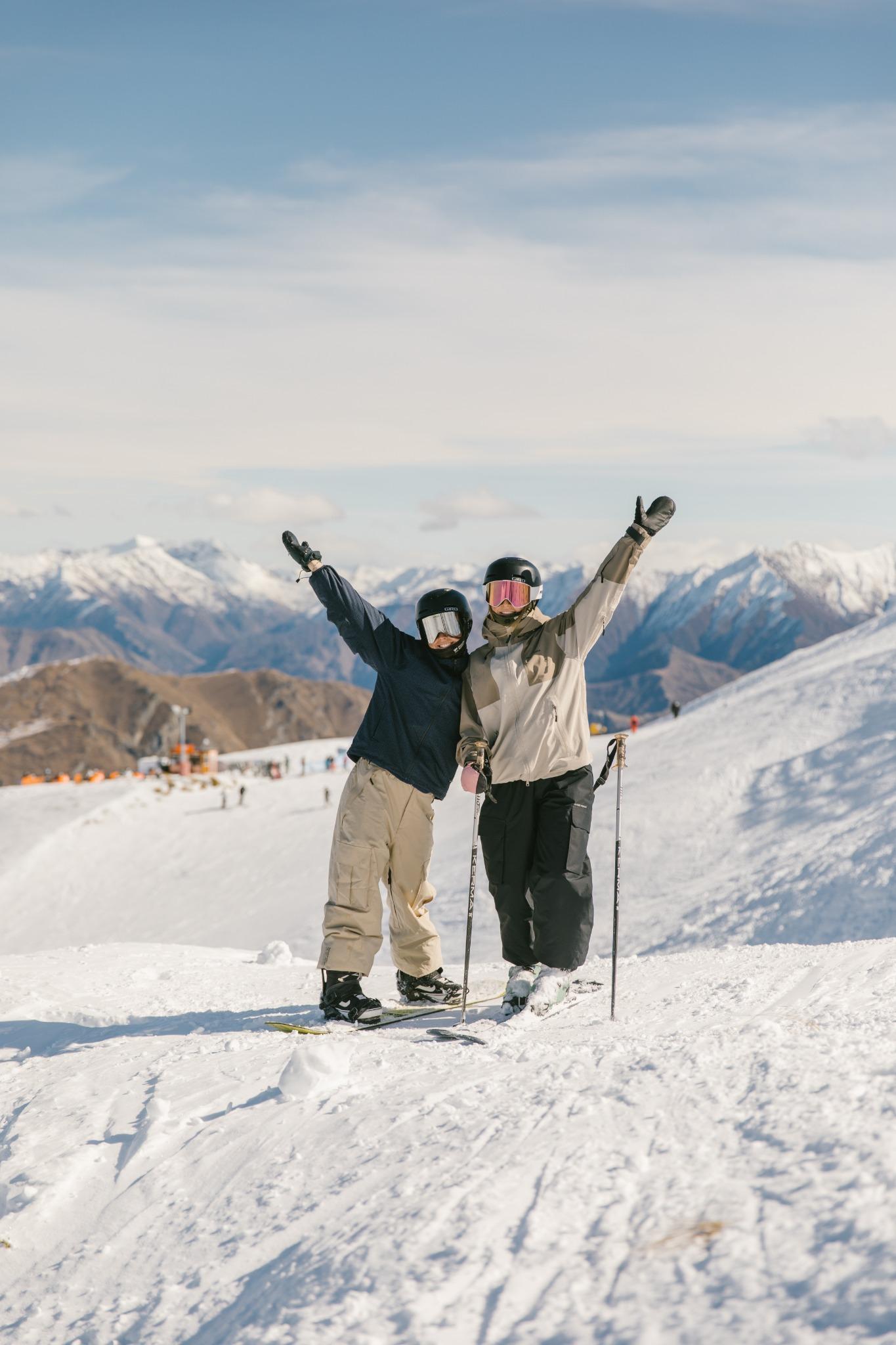 Two skiers wearing helmets and goggles raising arms on snowy slope with mountain range in background.