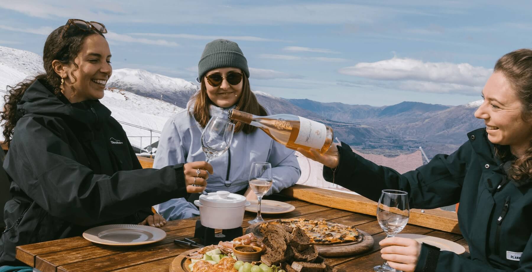 Three women in winter jackets share wine and food at a wooden table with snowy mountains behind.