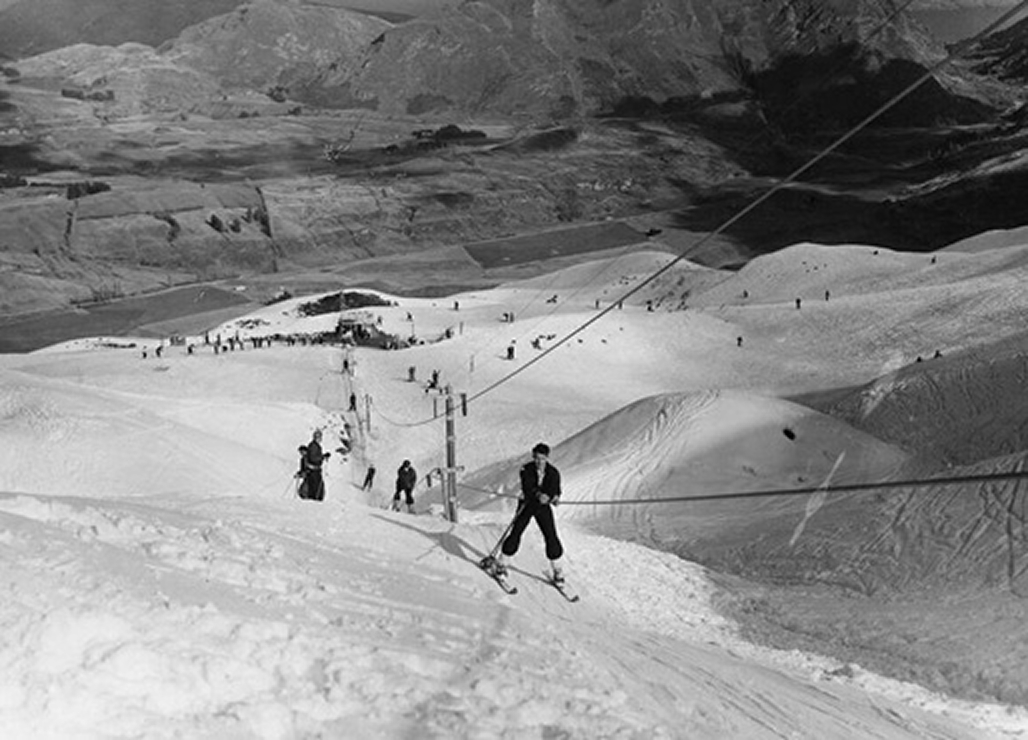 Black and white photo of skiers using a ski rope tow on a snowy mountain slope.