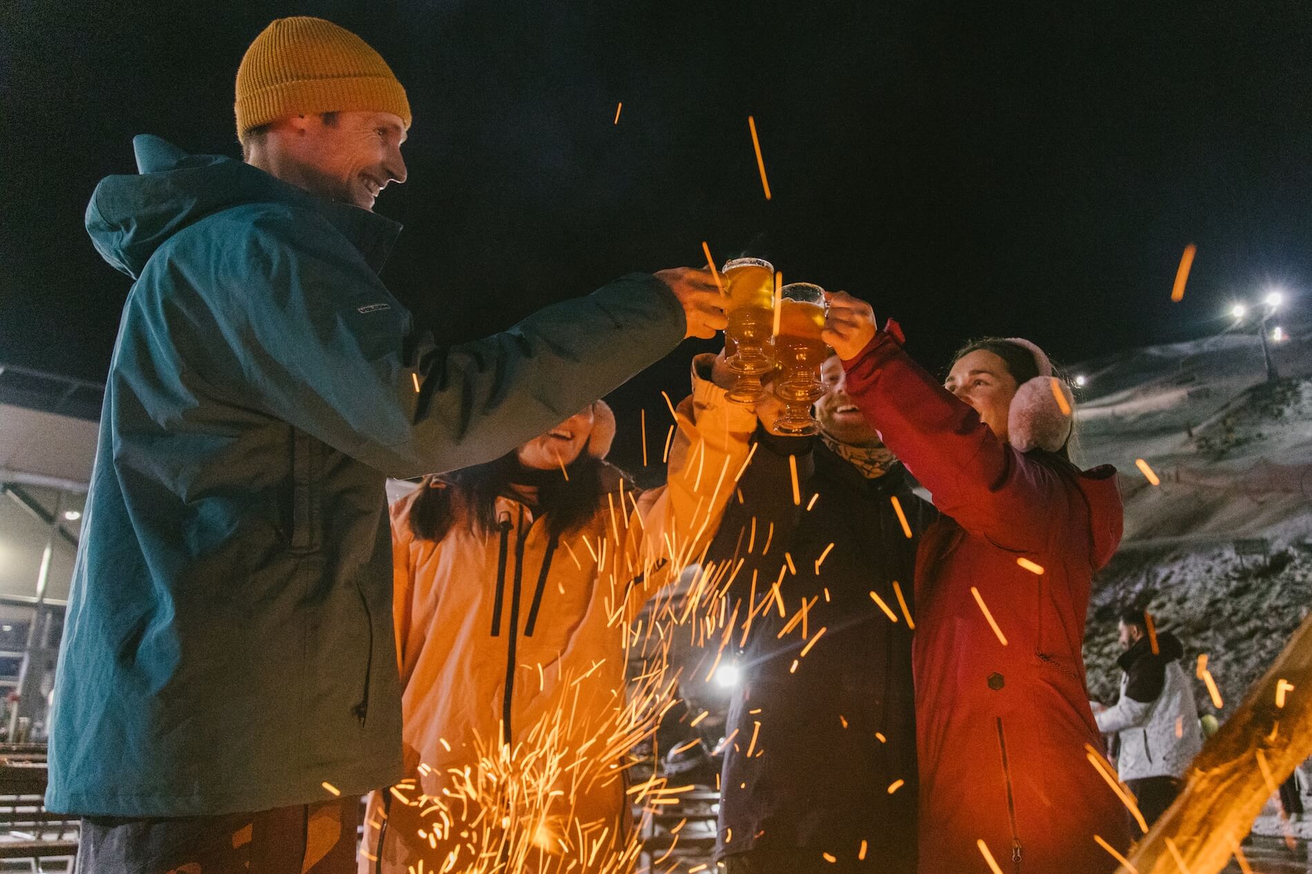 Group of four people toasting with drinks by a fire outdoors at night in winter clothes.