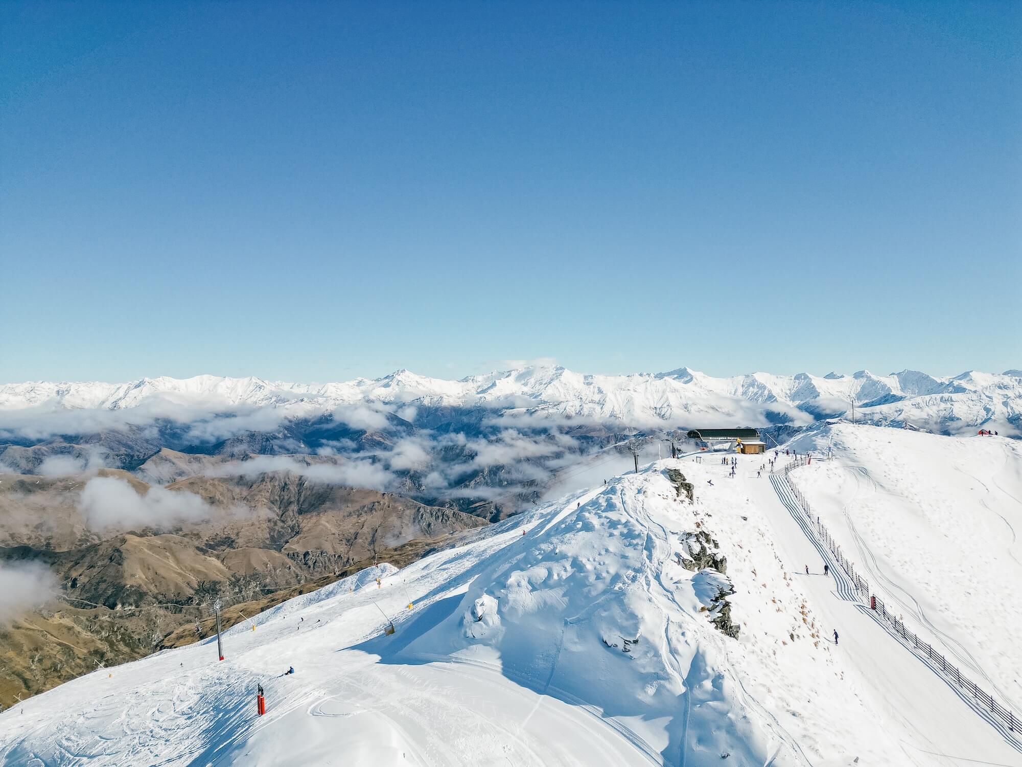 Snow-covered mountain peak with ski lift, skiers, and distant snowy mountain range under clear blue sky.