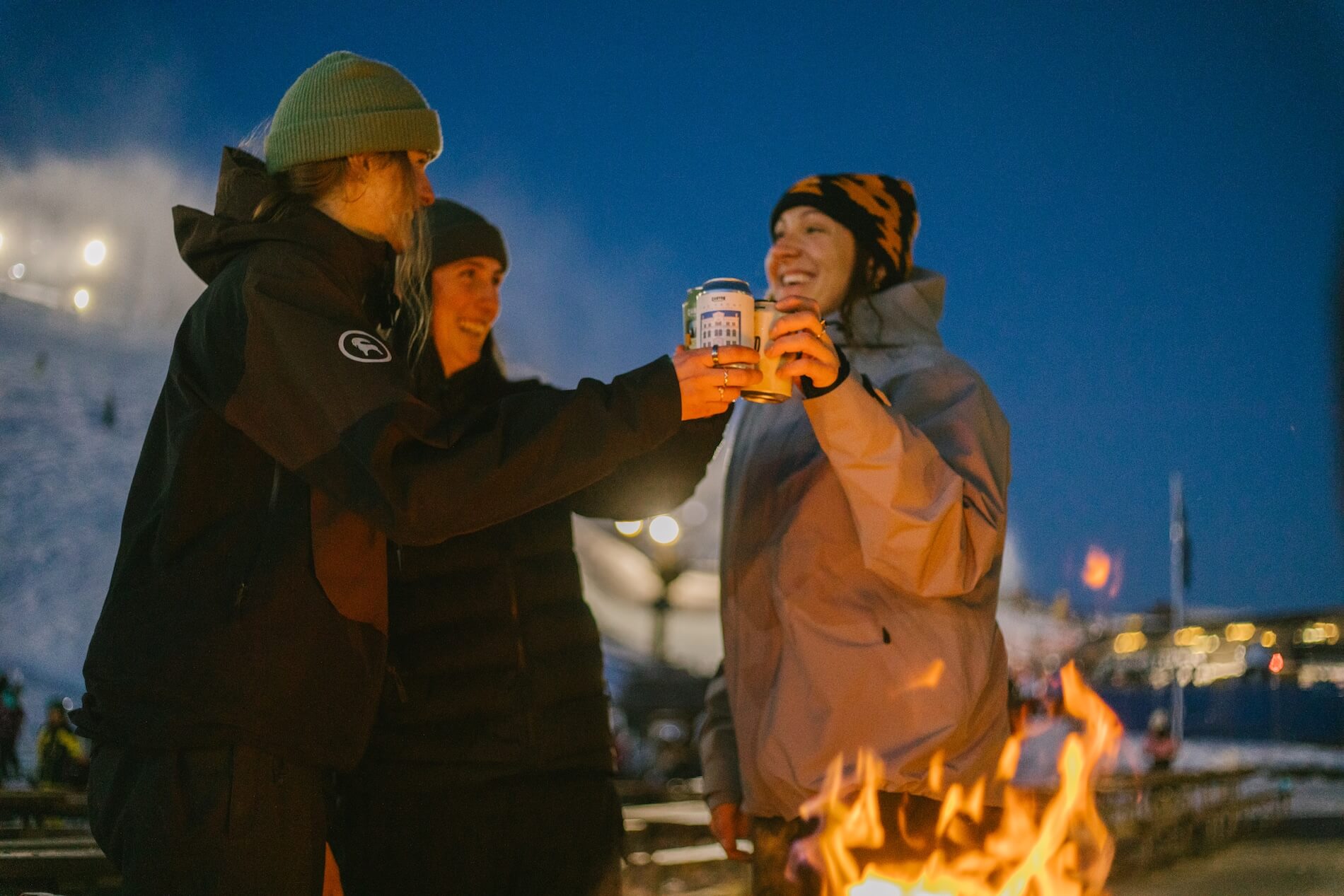 Three people in winter clothes toasting with drinks near a fire at dusk outdoors.