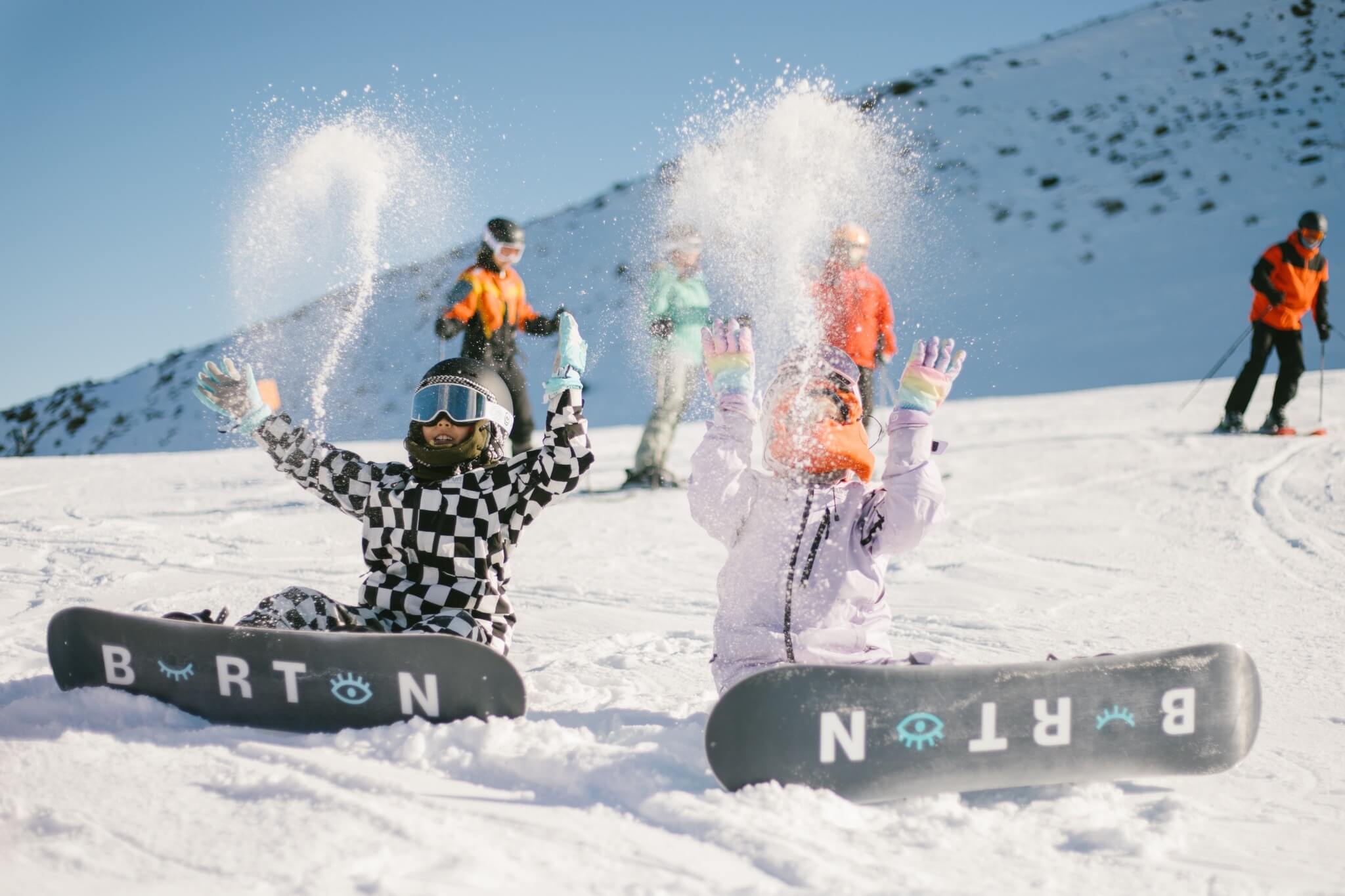 Two kids sitting on snow with snowboards, throwing snow up, and people skiing behind them.