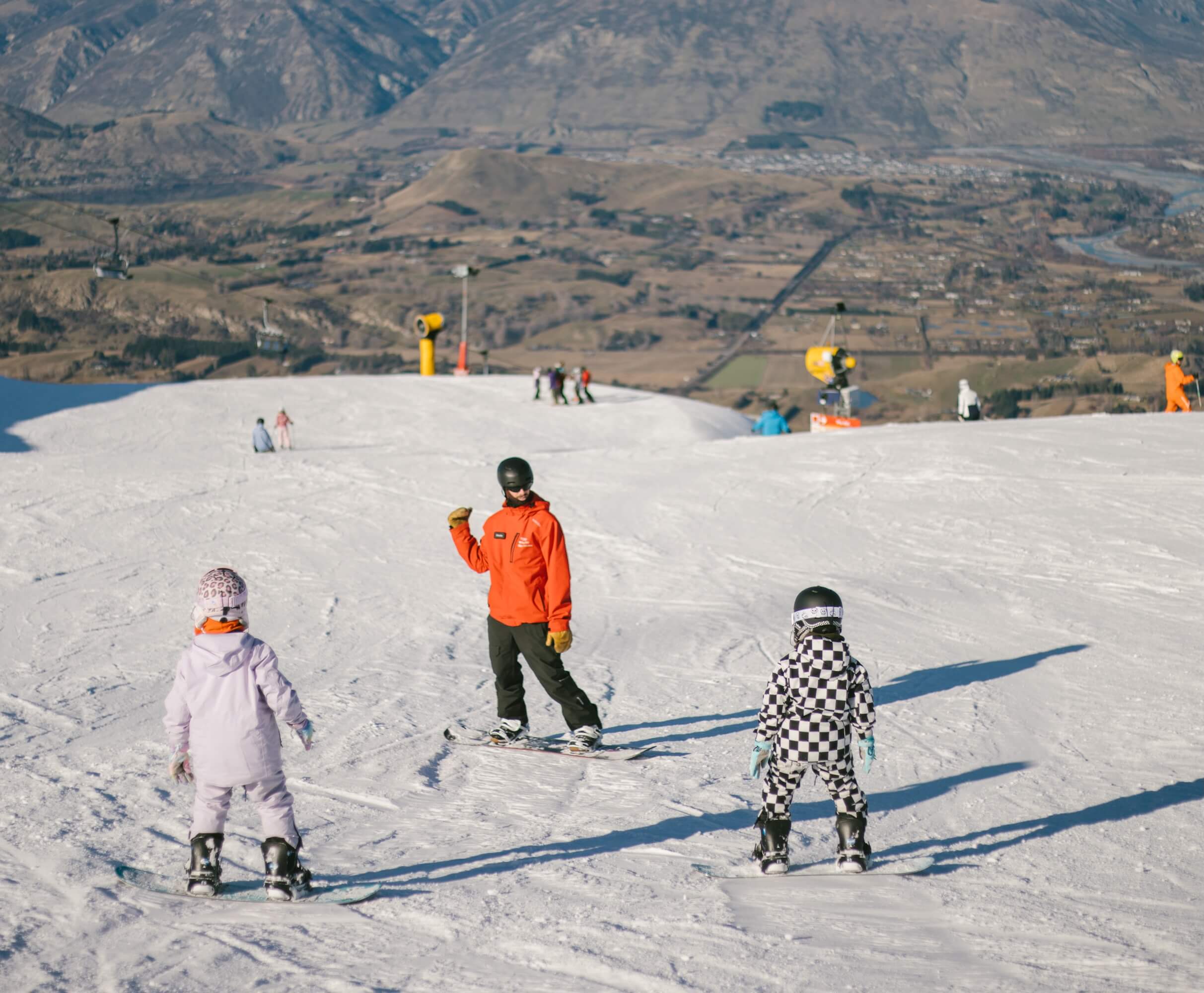 Ski instructor in orange jacket teaching two children on snow-covered slope with mountain valley background.
