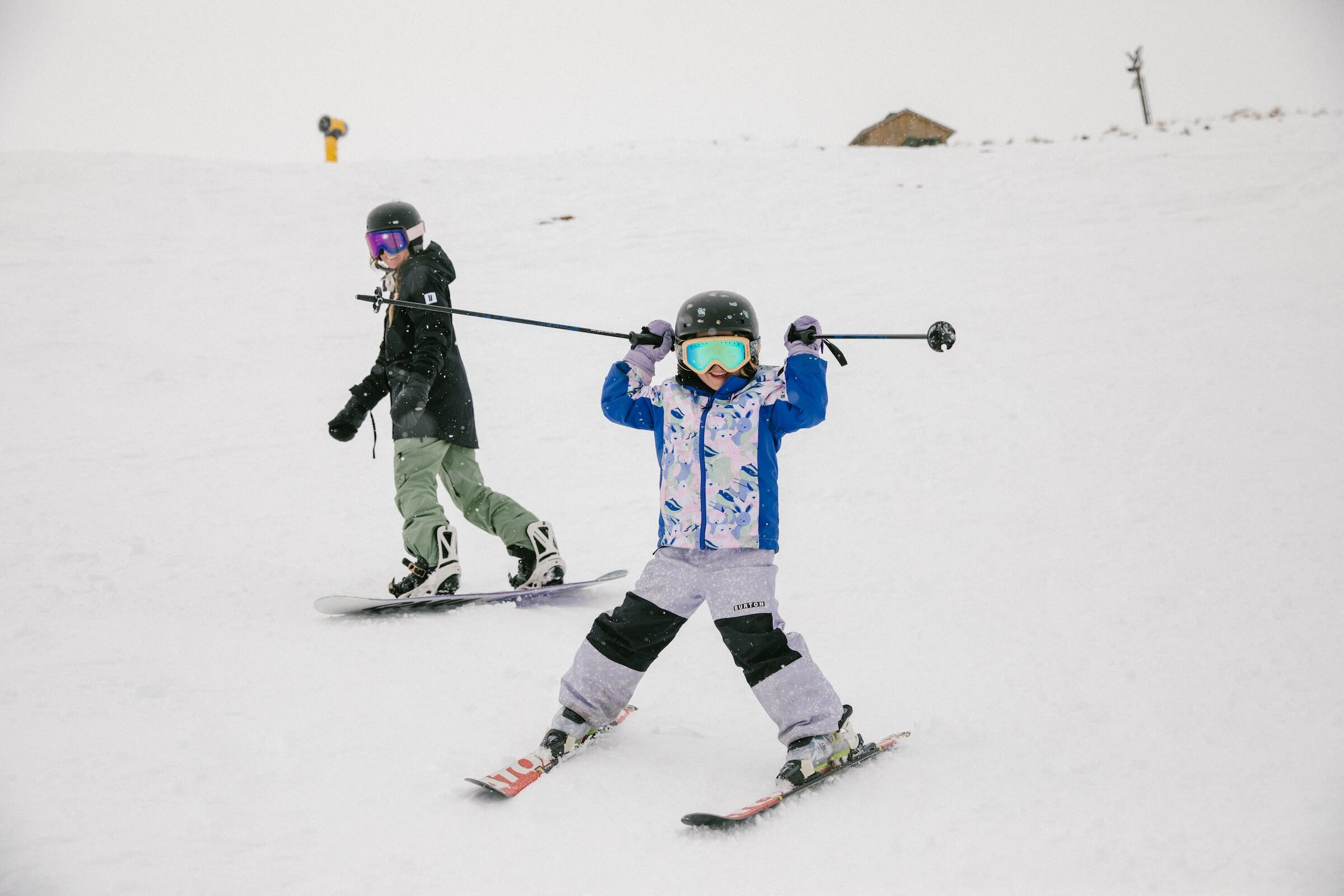 Child skiing with ski poles held overhead and an adult snowboarding on a snowy slope.