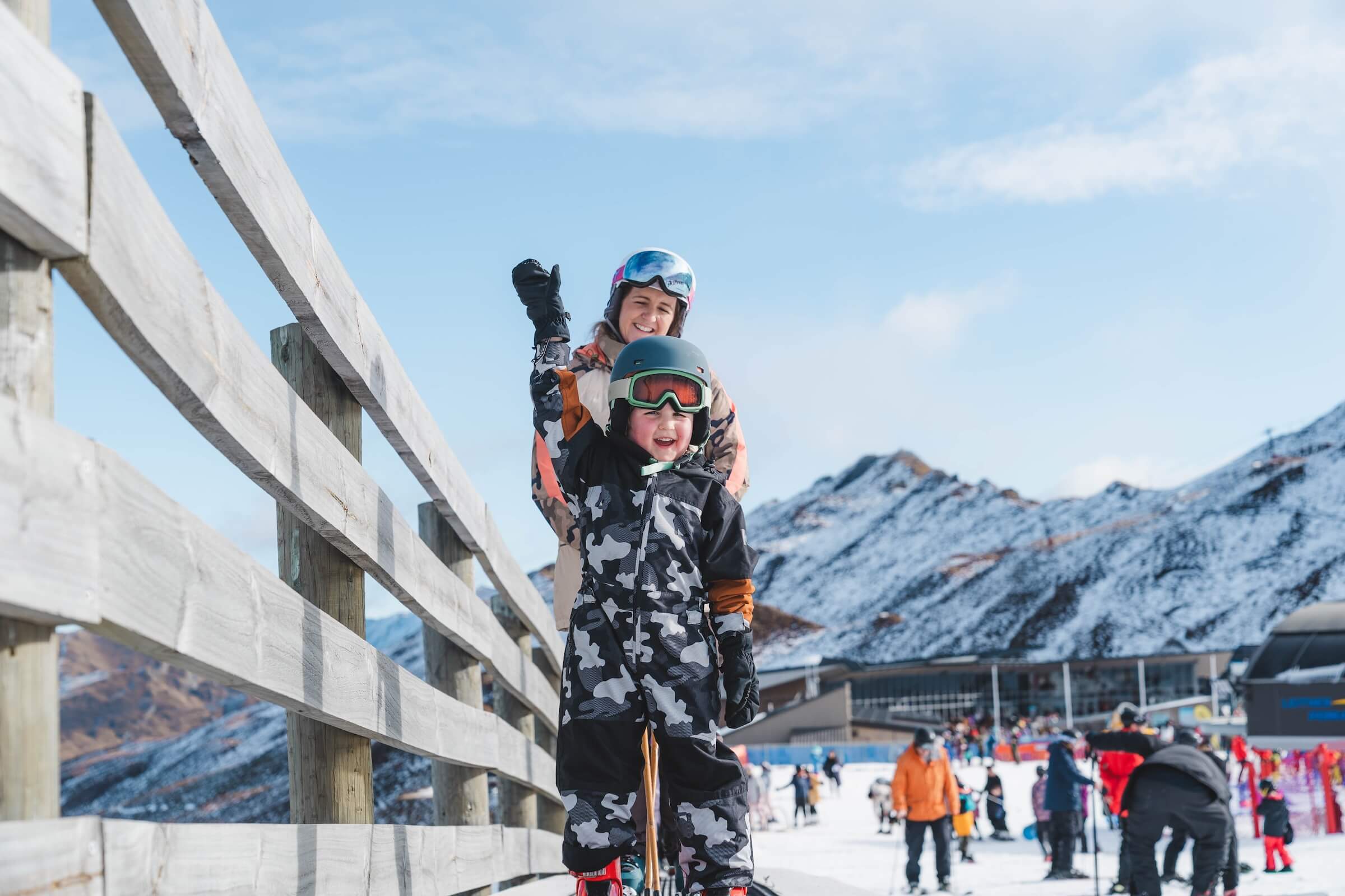 Child in camo ski suit with helmet and goggles raising arm, adult behind on snowy mountain ski resort.