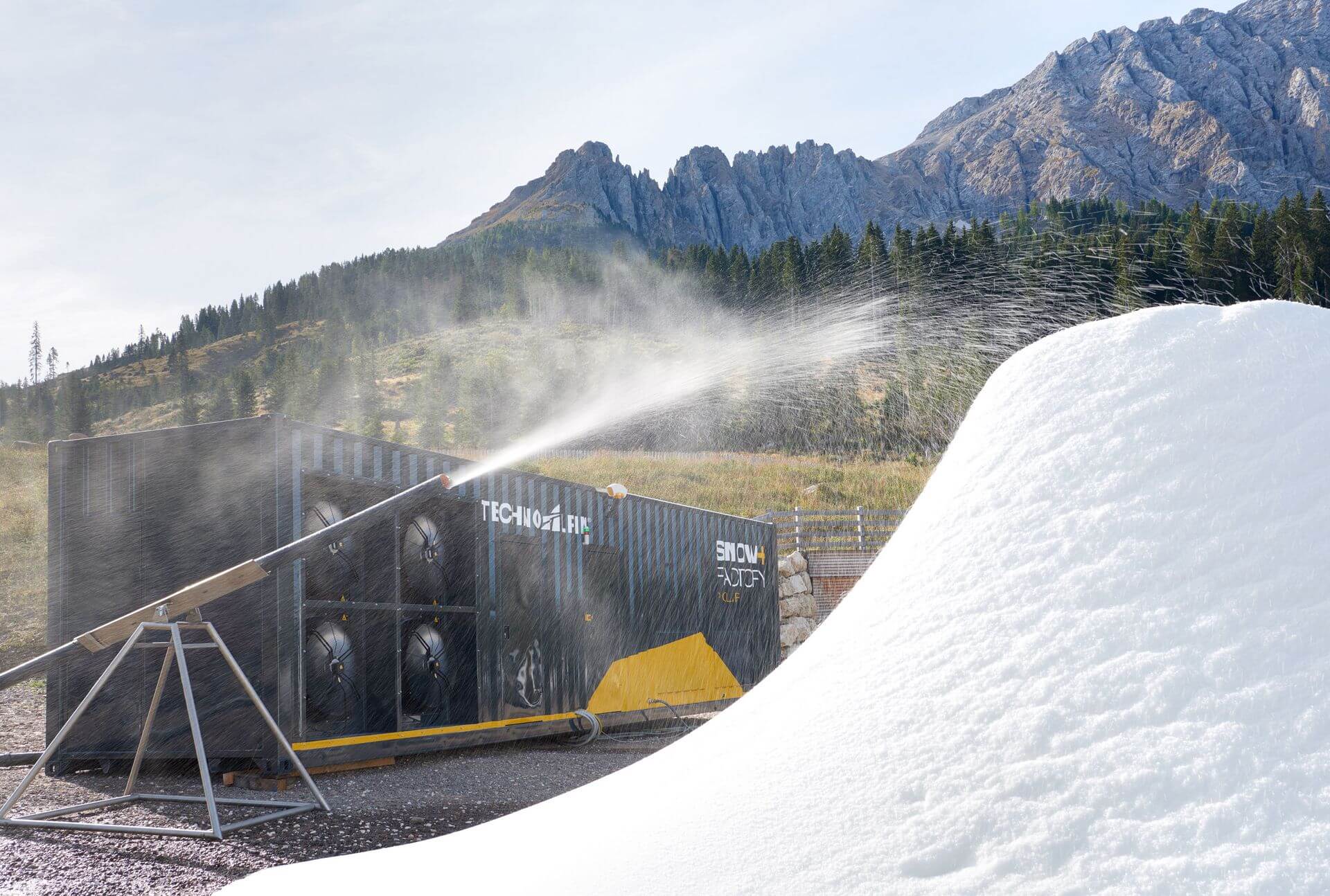 Snow machine spraying artificial snow onto a large mound with mountains and trees in the background.