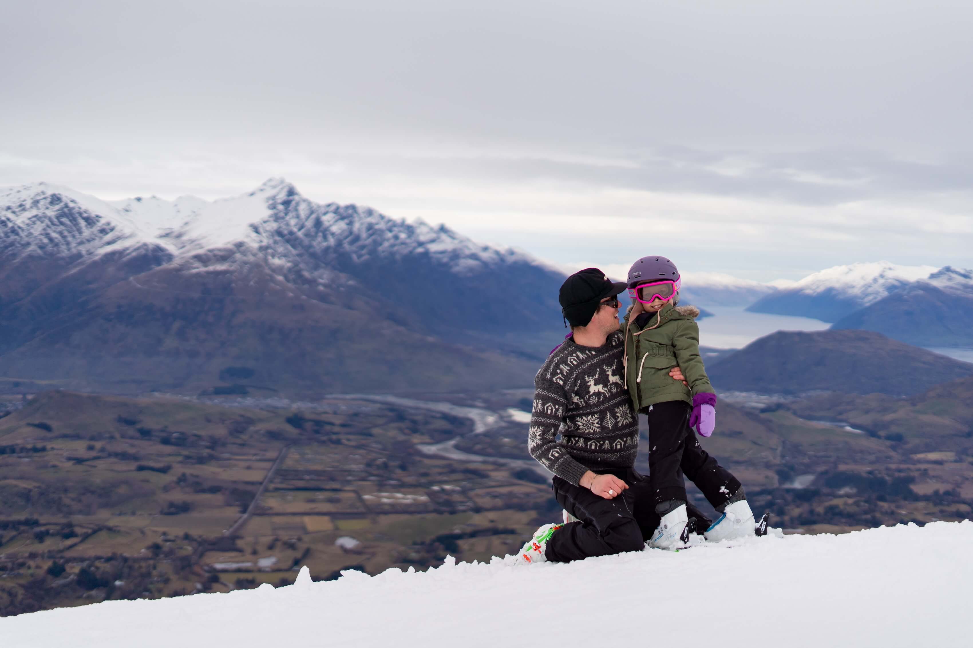 Man kneeling on snow holds a child in ski gear with snowy mountains and valley in the background.