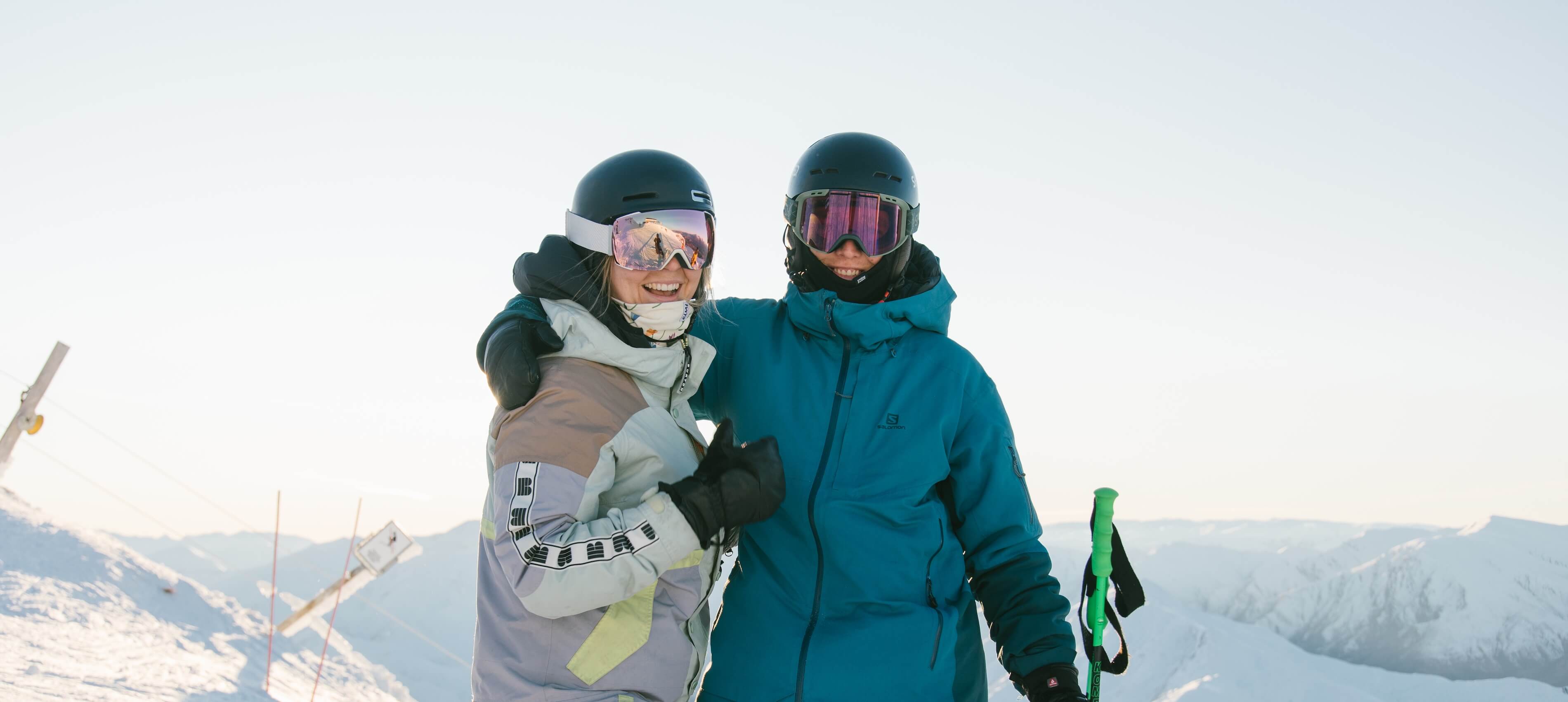 Two smiling skiers in winter gear and helmets posing together on a snowy mountain.
