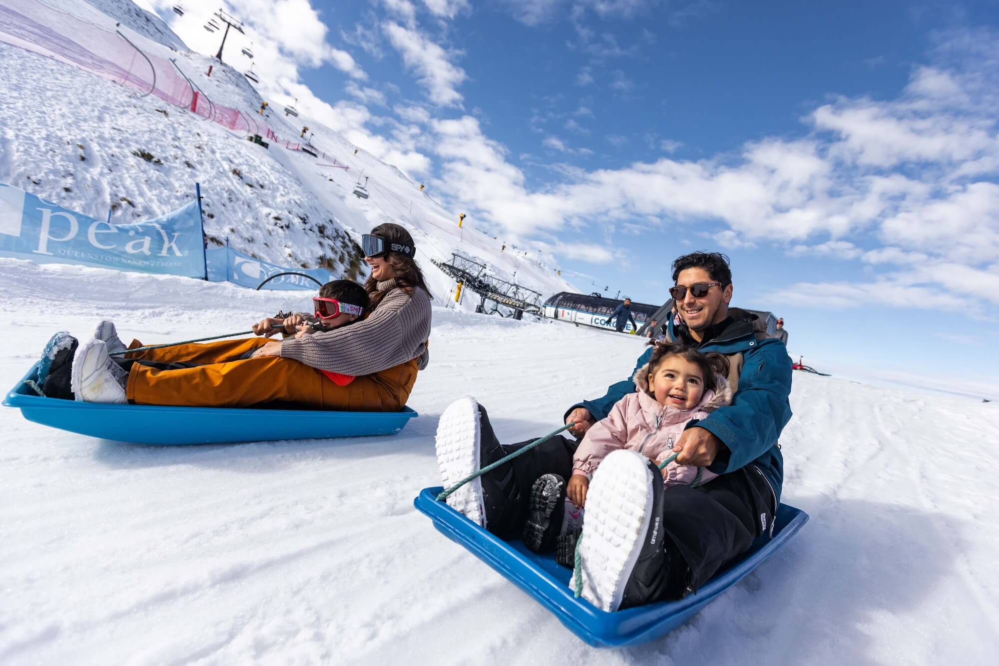 Two adults sledding down a snowy hill with children, all smiling and enjoying a sunny day.