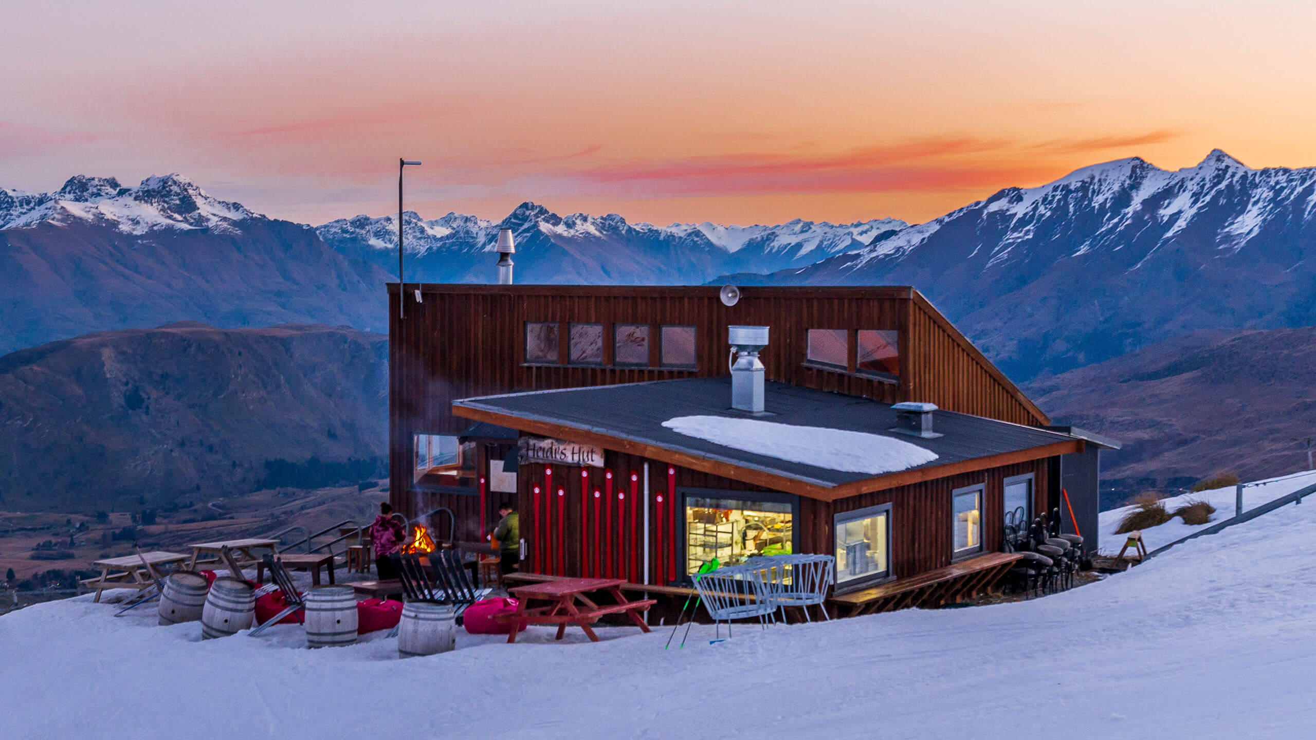 Wooden cabin with outdoor seating on snowy hill at sunset, with mountains in the background.