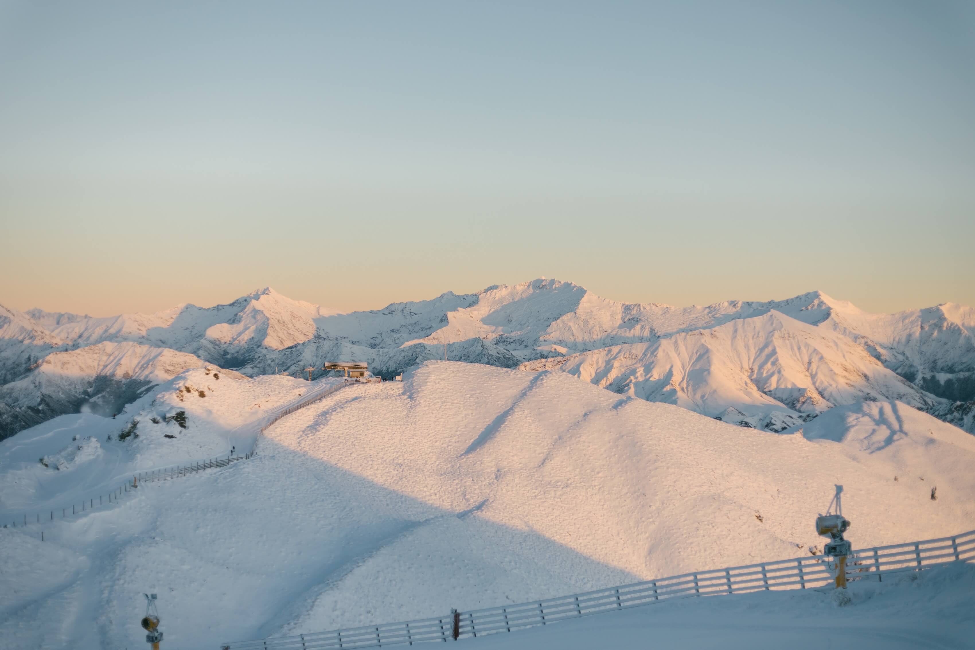 Snow-covered mountain range at sunrise with soft golden light and ski lift equipment in foreground.