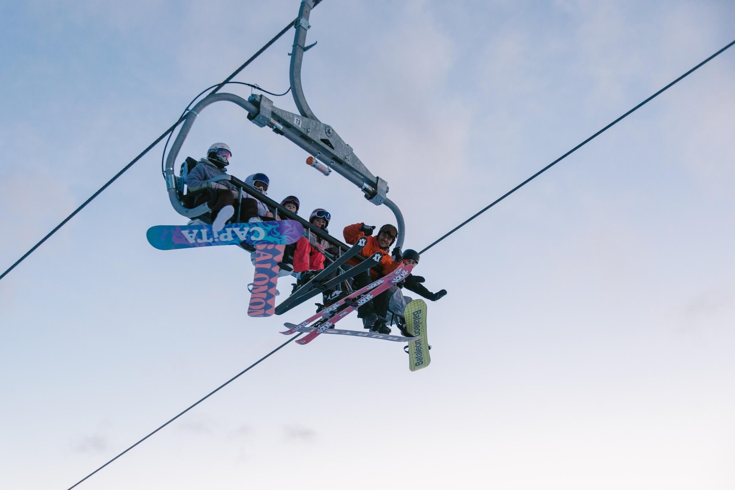 Four people wearing ski gear riding a ski lift with snowboards and skis suspended in the air.