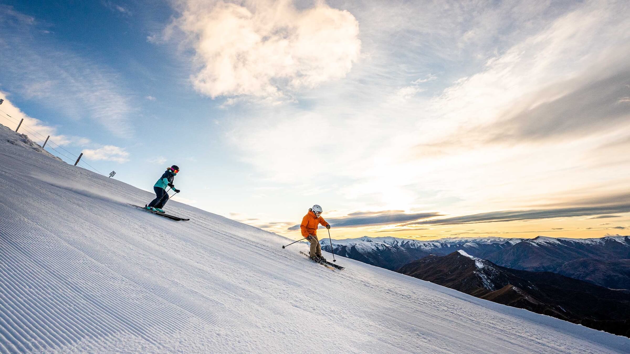 Two skiers descending a snowy slope with mountains and a cloudy sky at sunset.