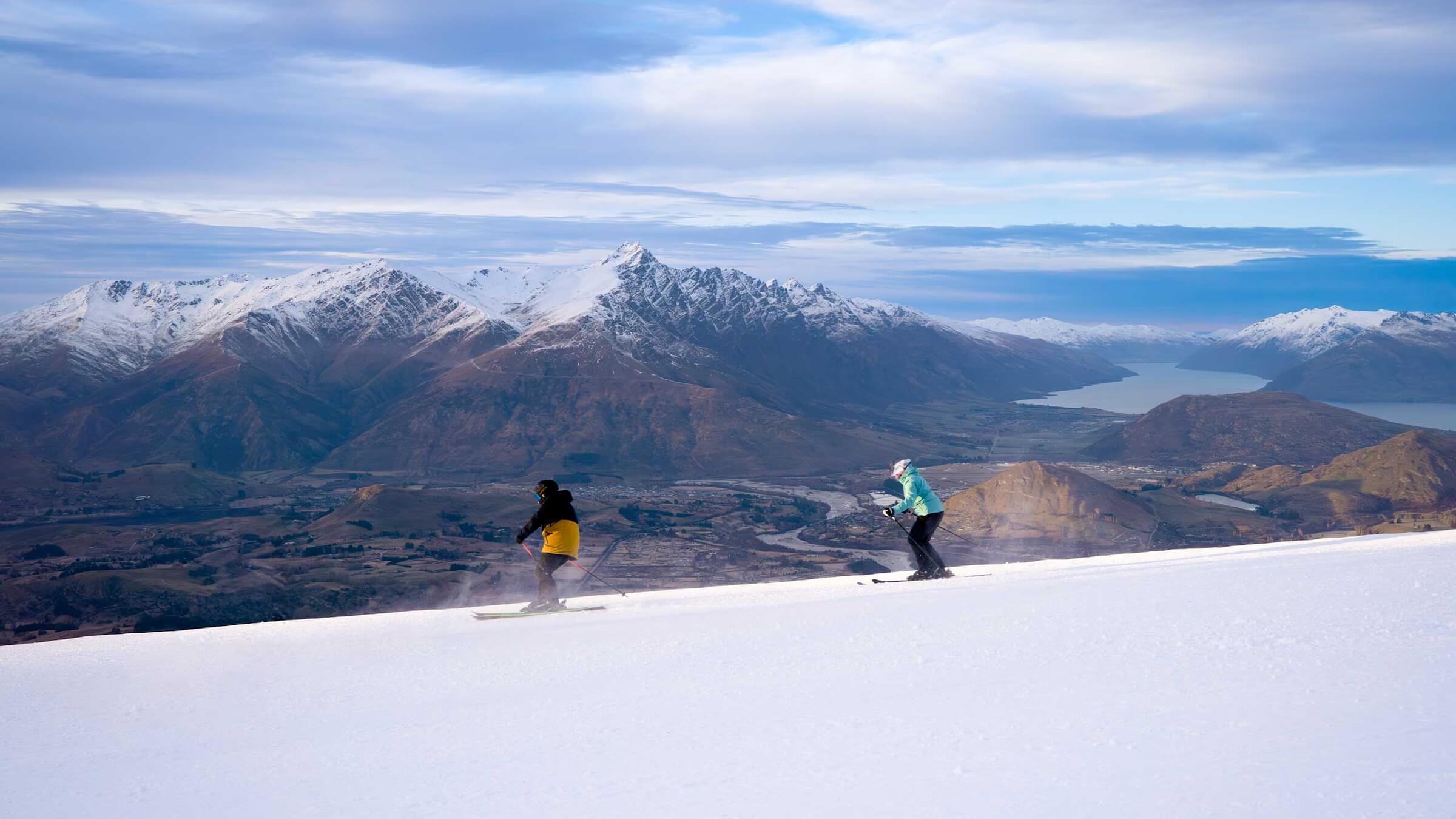 Two skiers descend a snowy slope with snow-capped mountains and a lake in the distance.