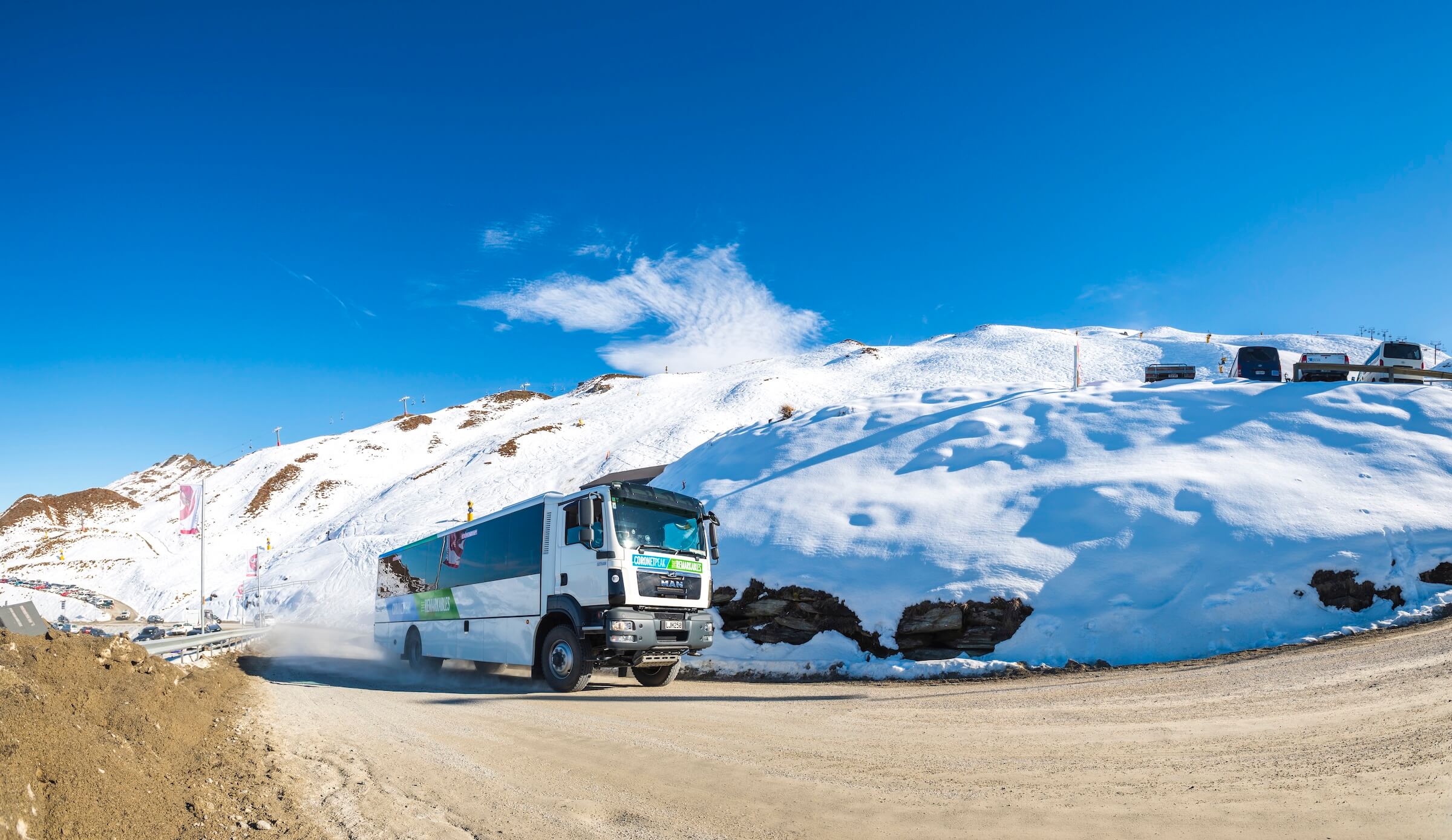 White bus with green signs drives on a dirt road near snowy mountains under a clear blue sky.