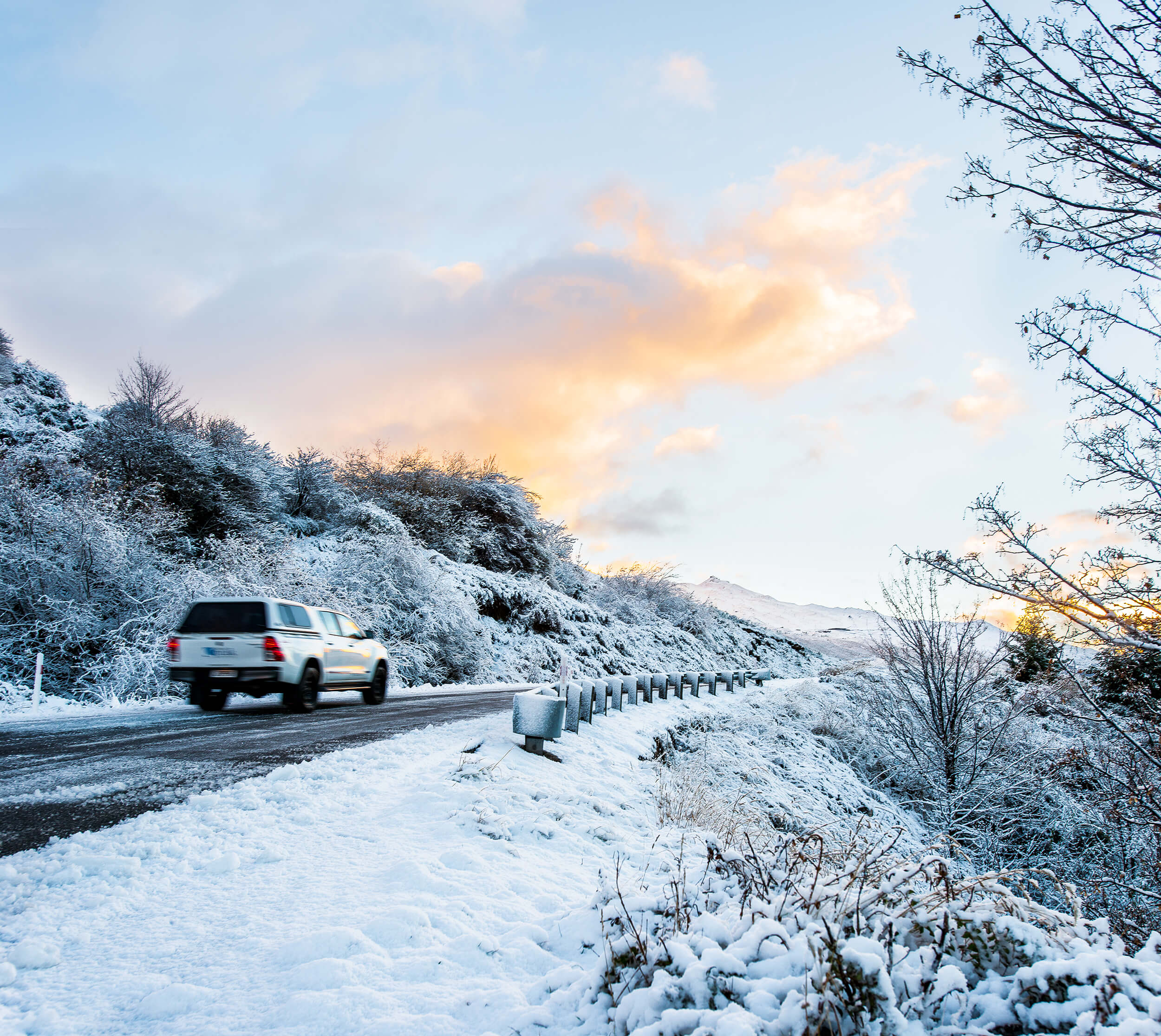 White SUV driving on a snowy road lined with snow-covered trees under a partly cloudy sky at sunset.