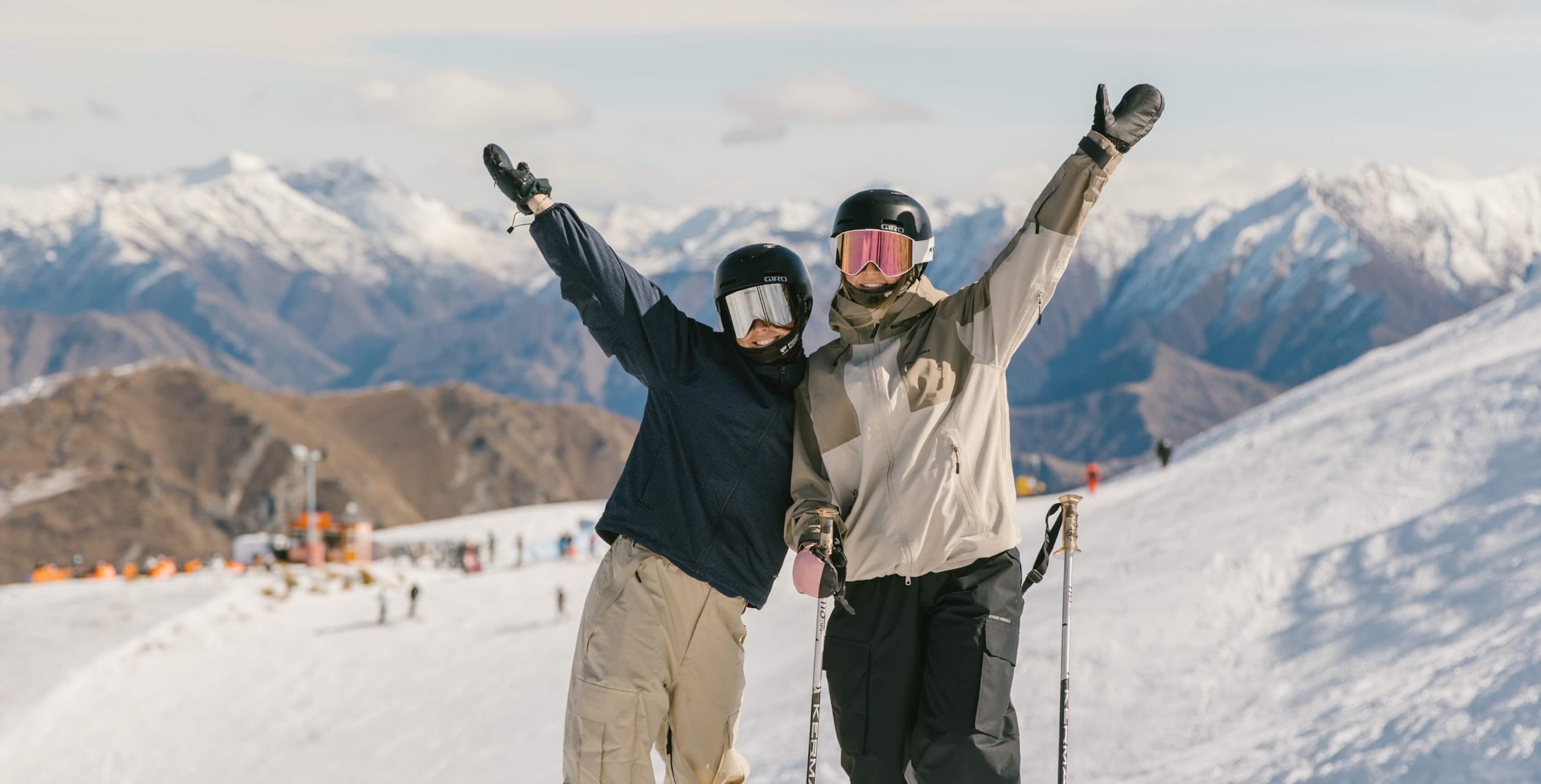 Two skiers wearing helmets and goggles pose with arms raised on a snowy mountain slope.