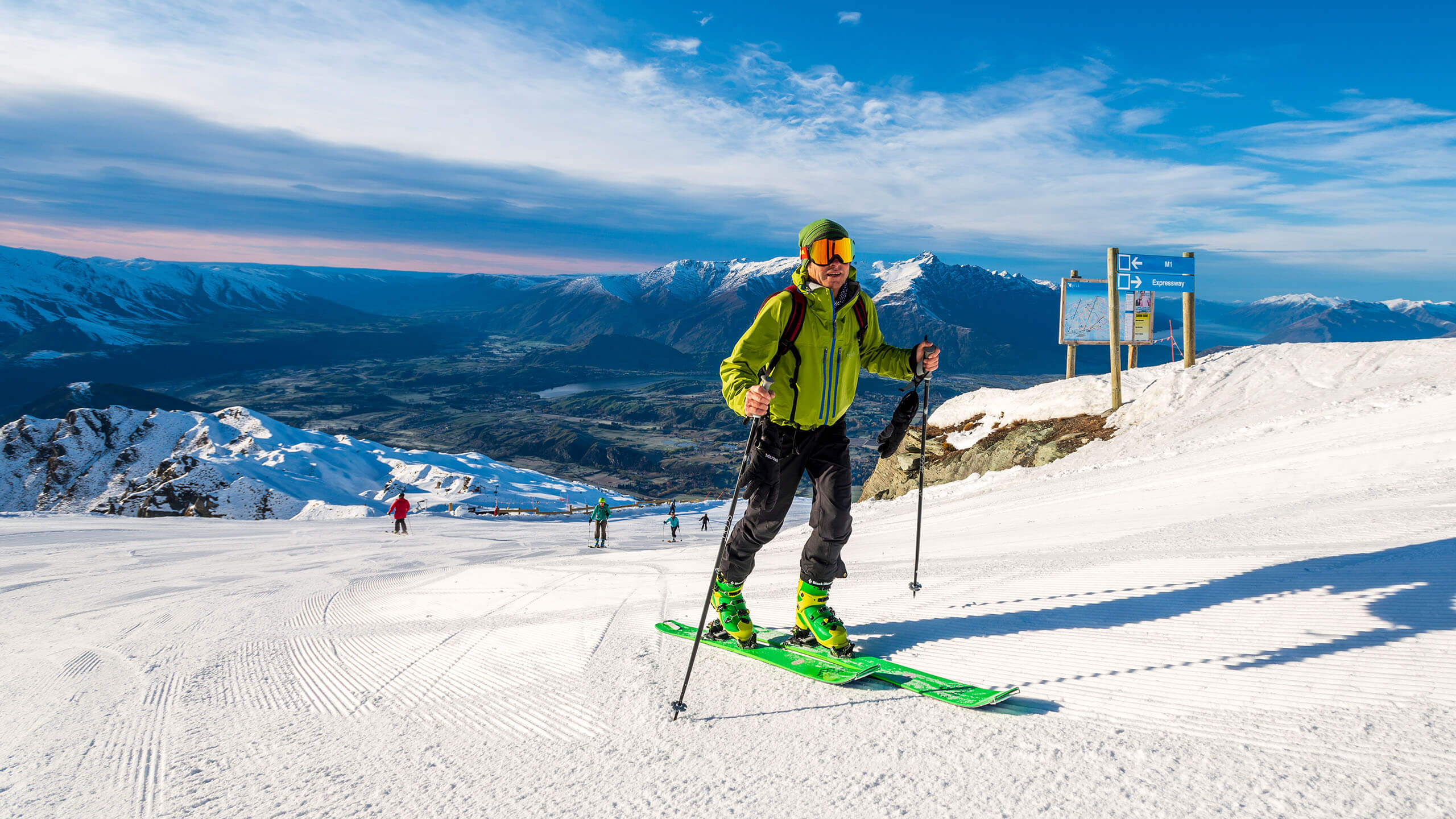 Skier in a green jacket and skis on a snowy slope with mountains and blue sky behind.
