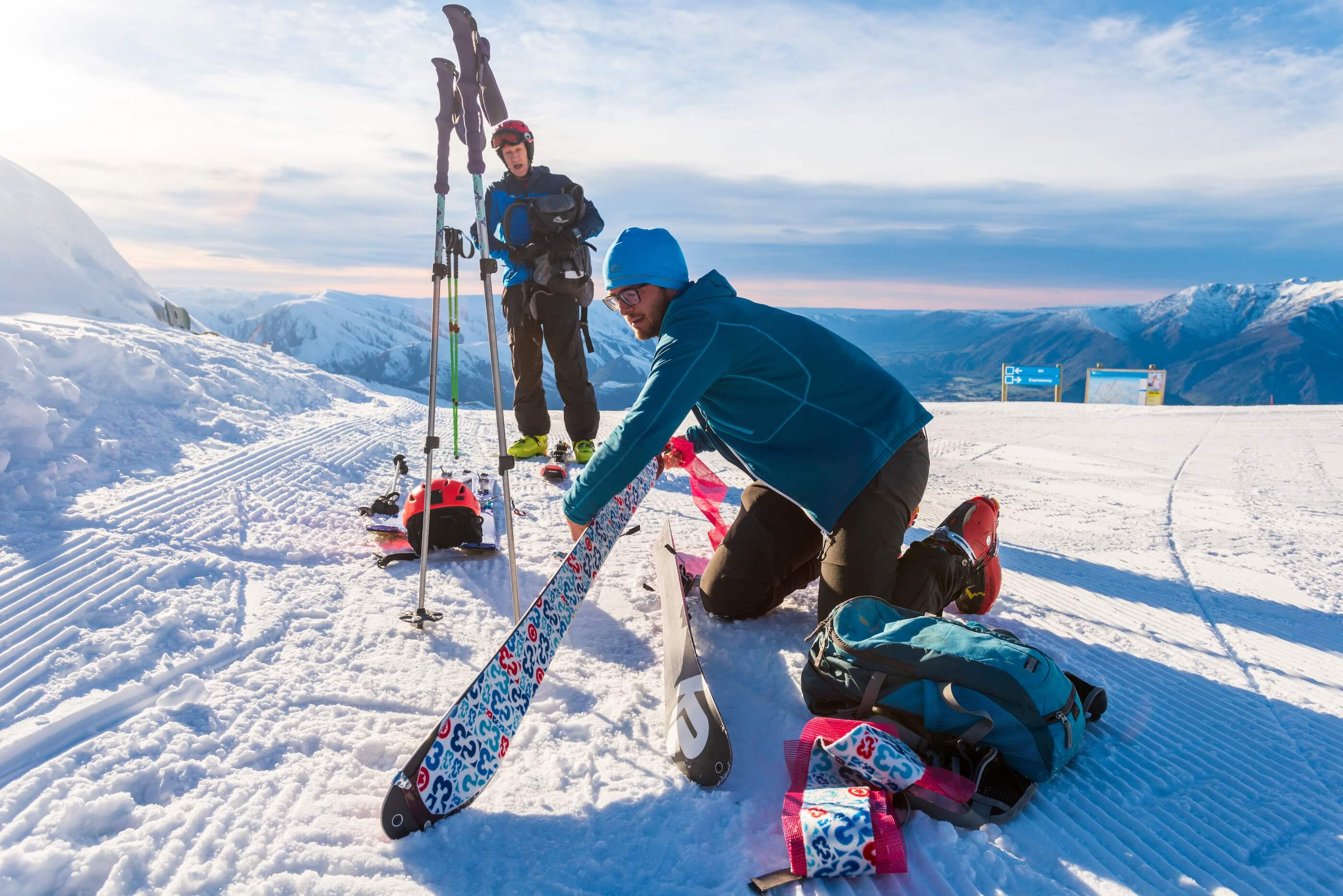 Two skiers on snowy slope, one kneeling adjusting skis, mountains and blue sky in background.