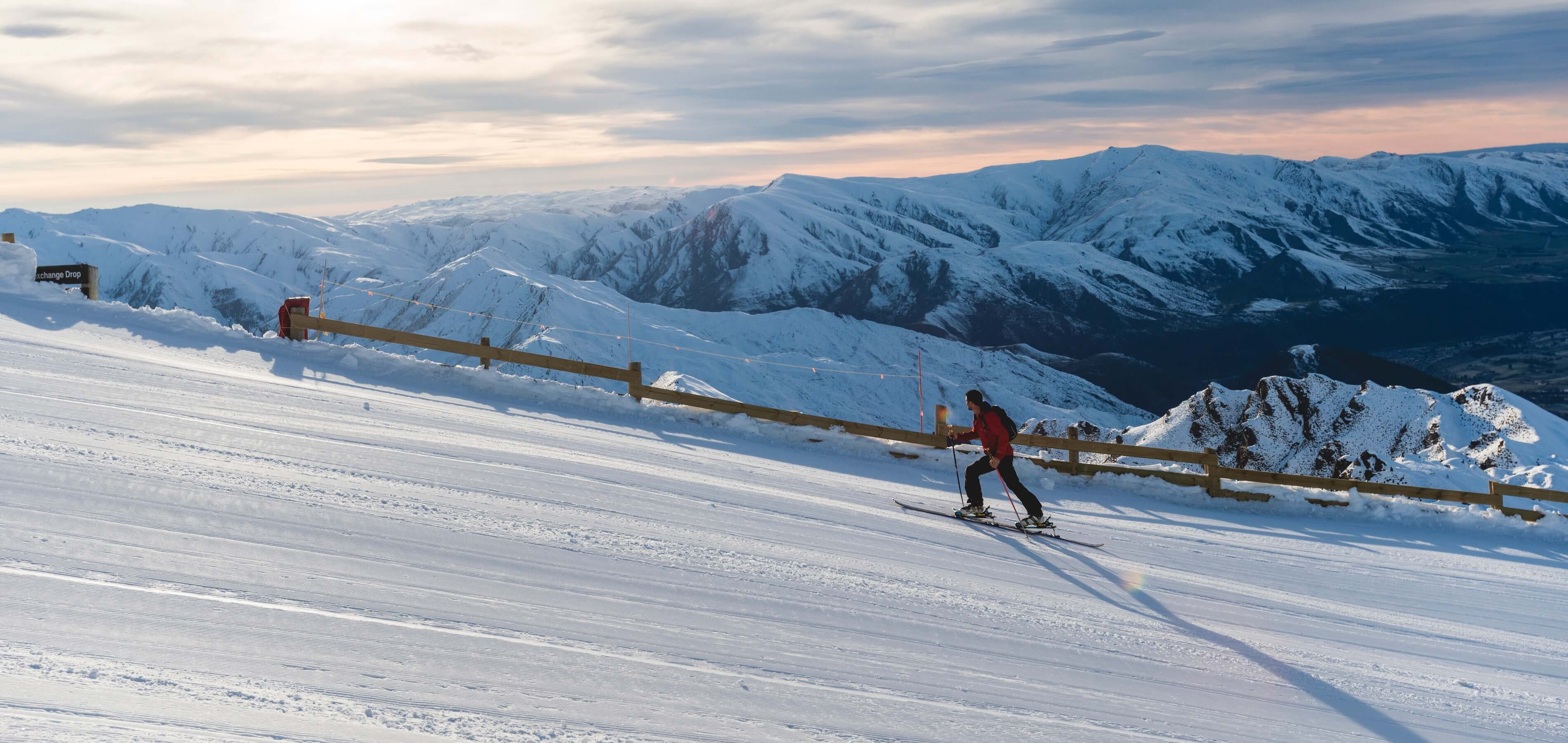 Skier in red jacket skiing downhill on a snowy mountain slope with distant snow-covered peaks.