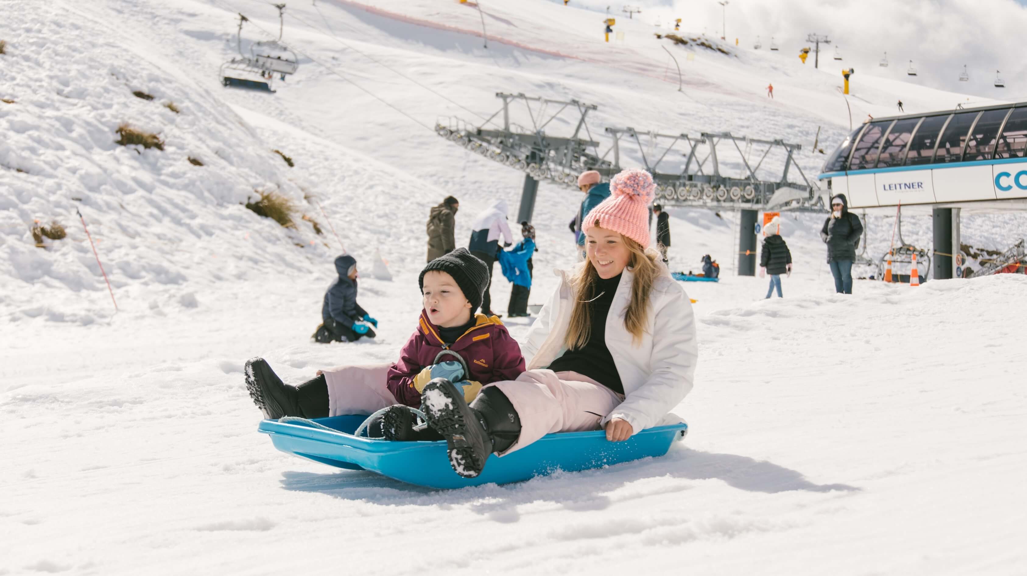 A woman and child sledding on snow at a ski resort with chairlifts and other people in the background.