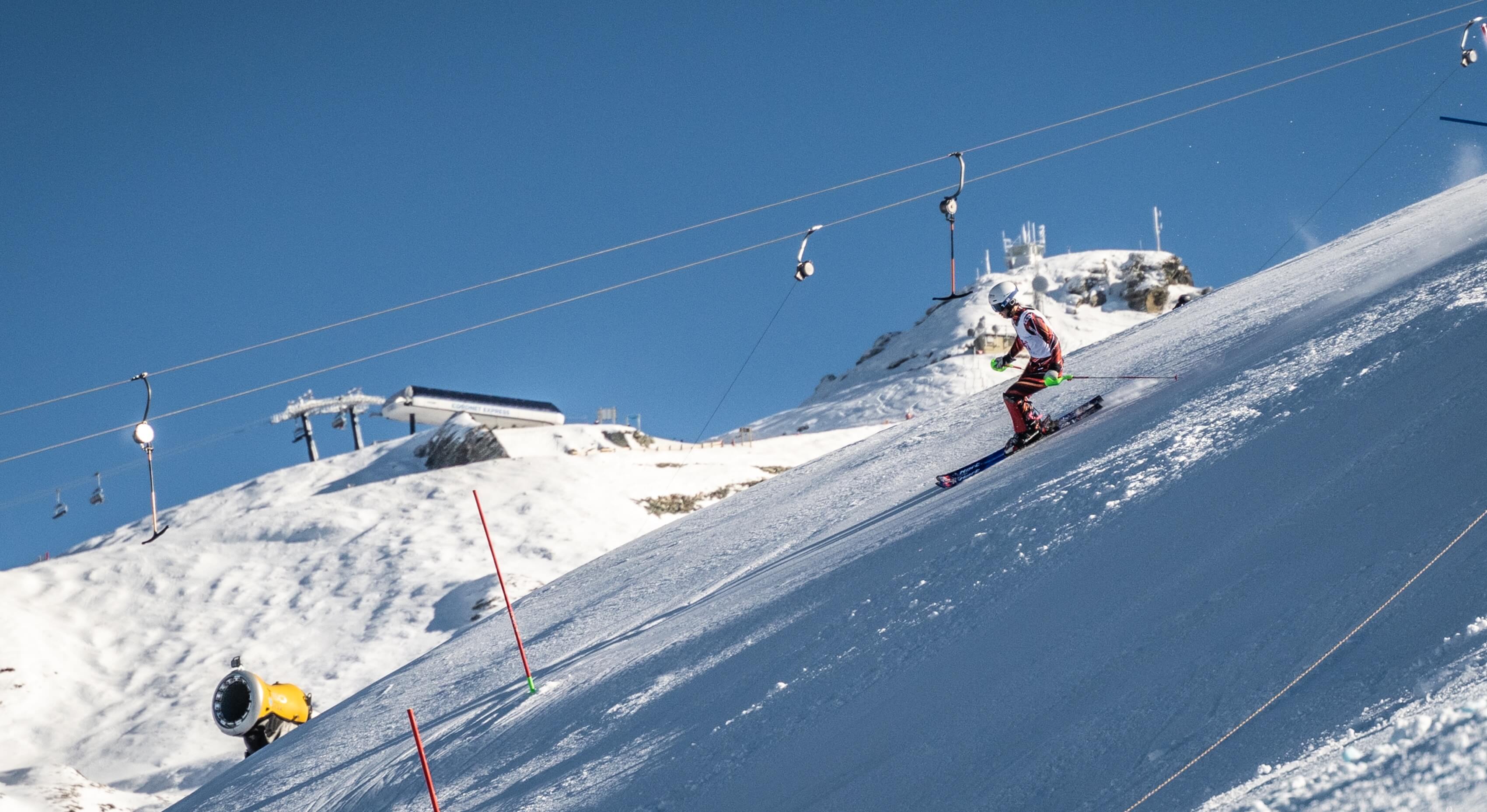Skier in racing gear skiing down snowy slope near ski lift and snowmaking machine.