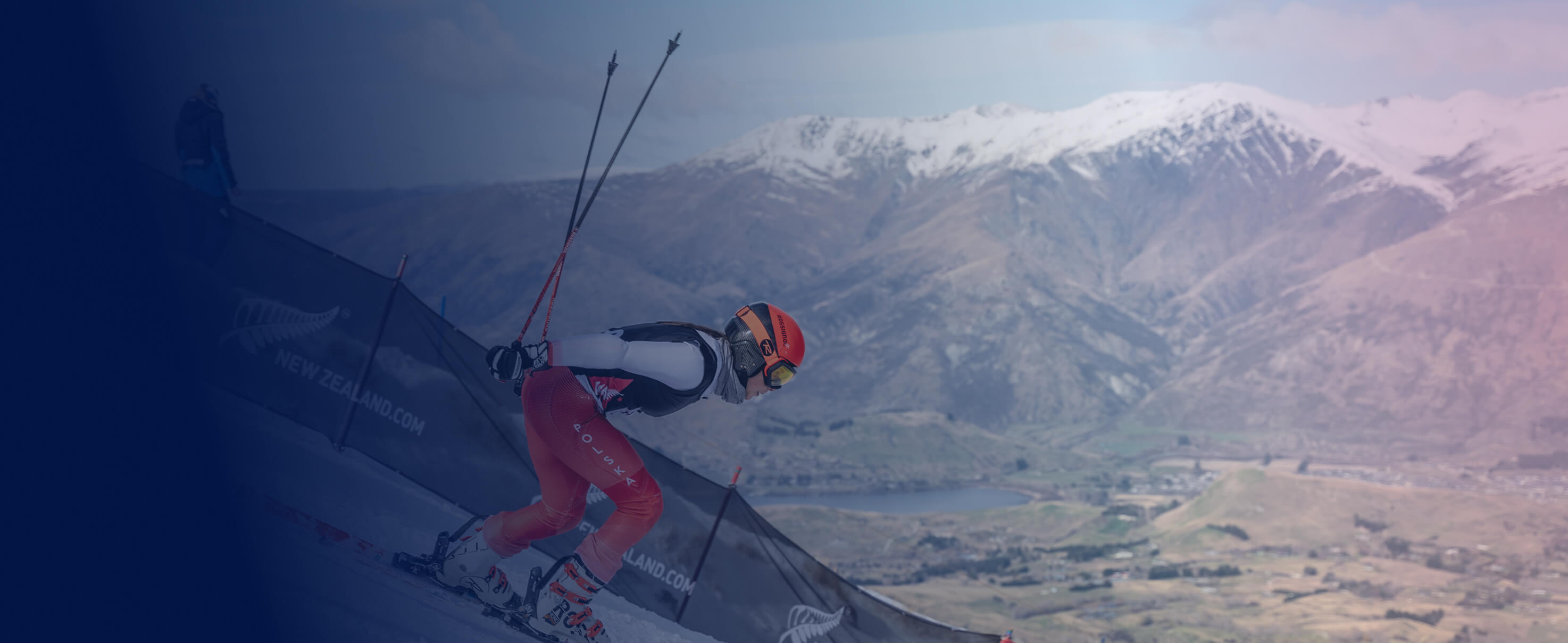 Skier in red gear and helmet racing downhill with snow-capped mountains and valley in the background.
