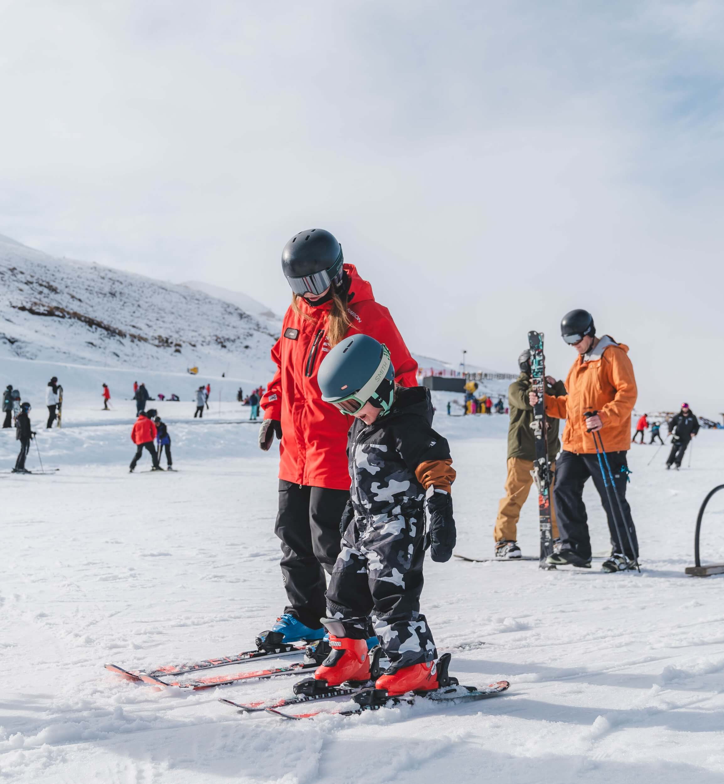 Ski instructor in red teaching child with helmet and camo suit to ski on snowy slope.