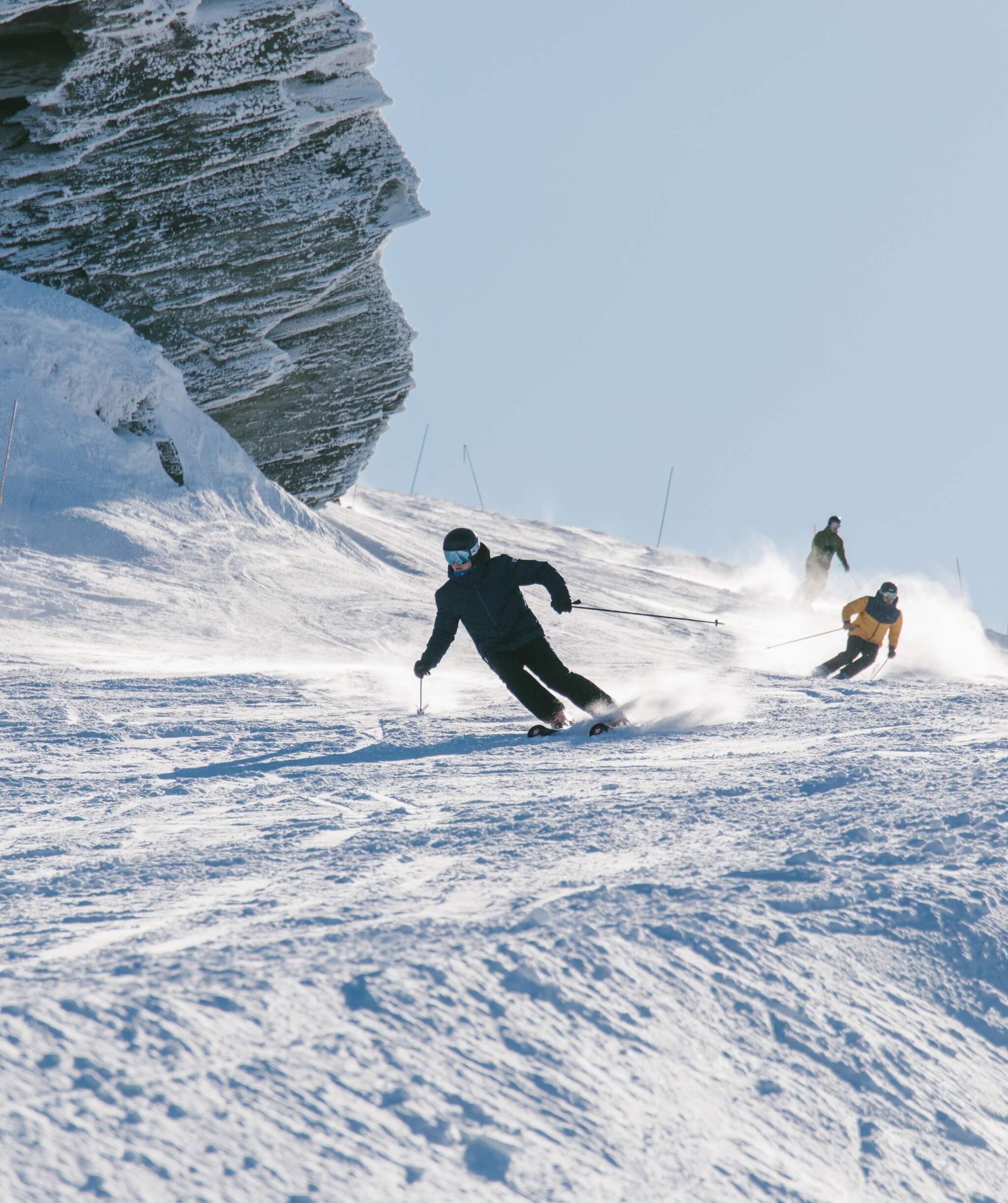 Three skiers descend a snowy slope near a rocky, snow-covered mountain on a clear day.