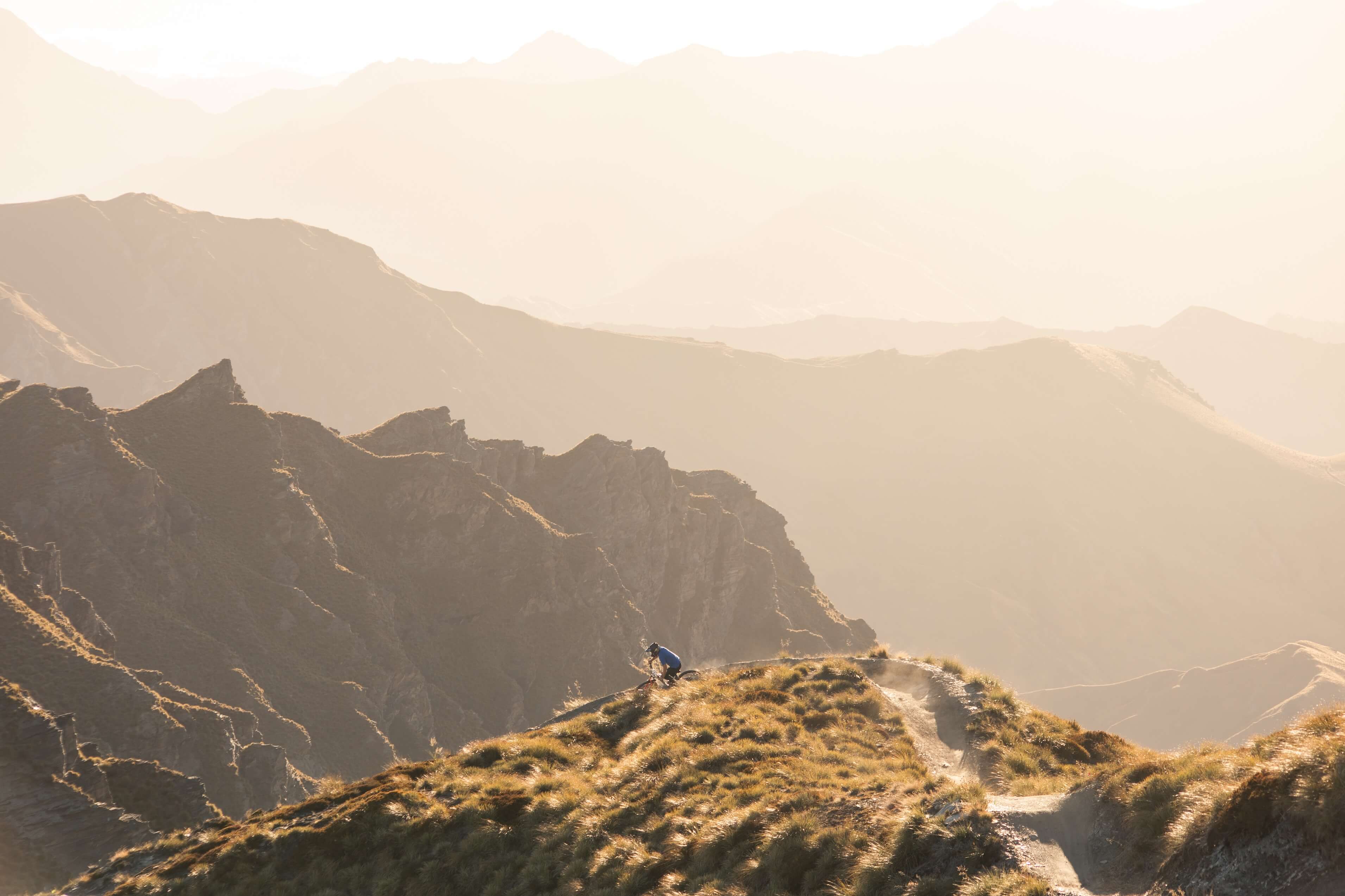 Mountain biker in blue rides on narrow dirt trail atop a grassy ridge with hazy mountains behind.