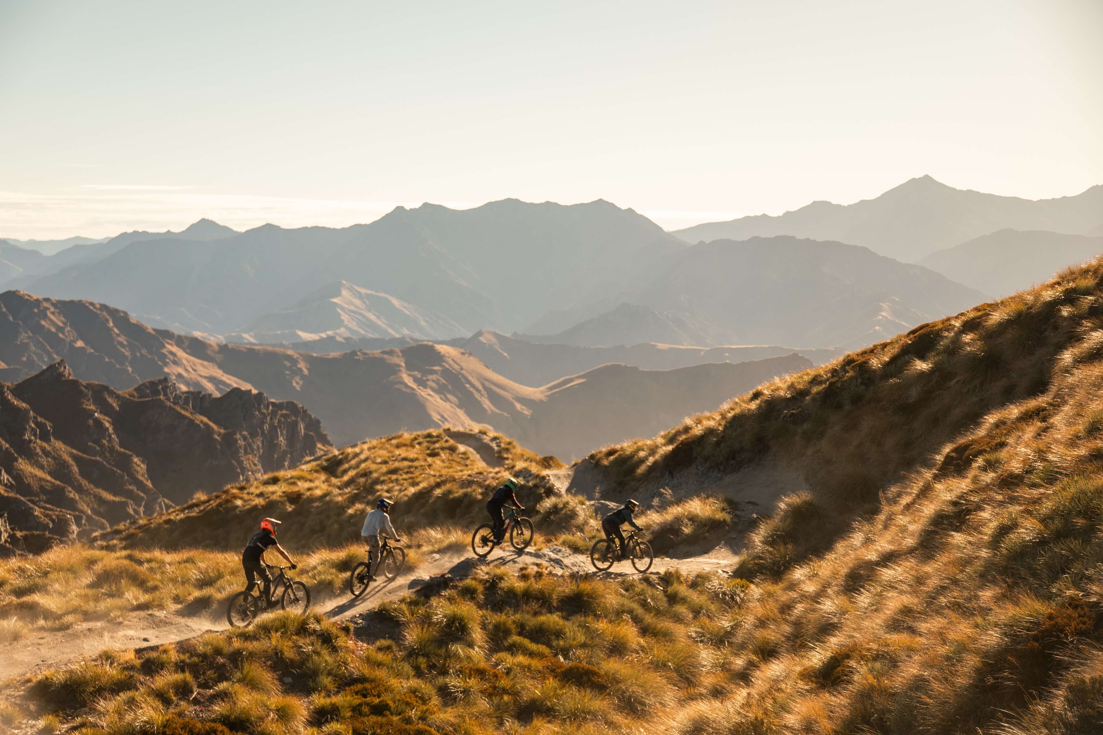 Four mountain bikers ride a trail along grassy hills with distant mountain ranges under clear sky.