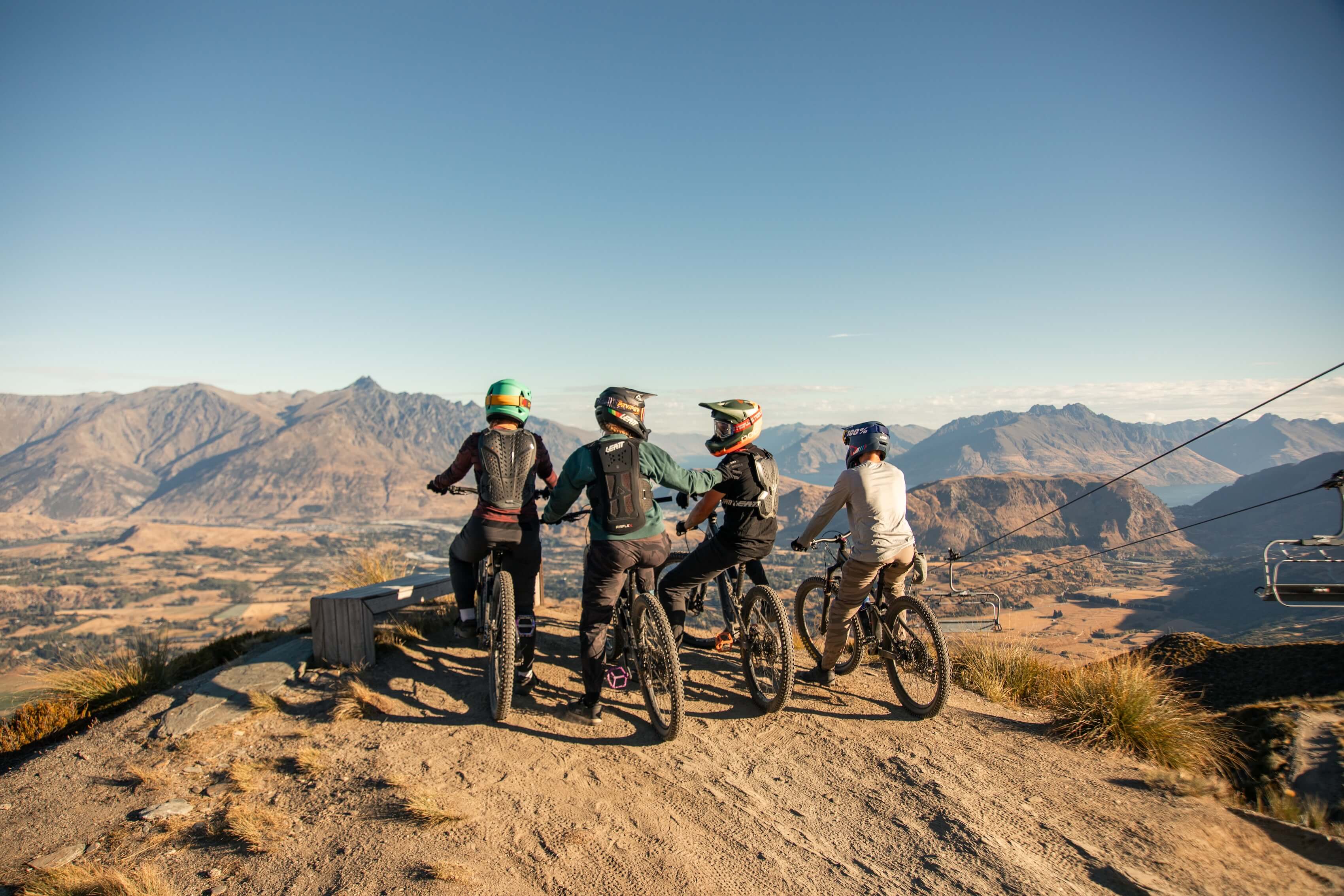 Four mountain bikers wearing helmets and gear pause at a high trail overlooking rugged mountains.