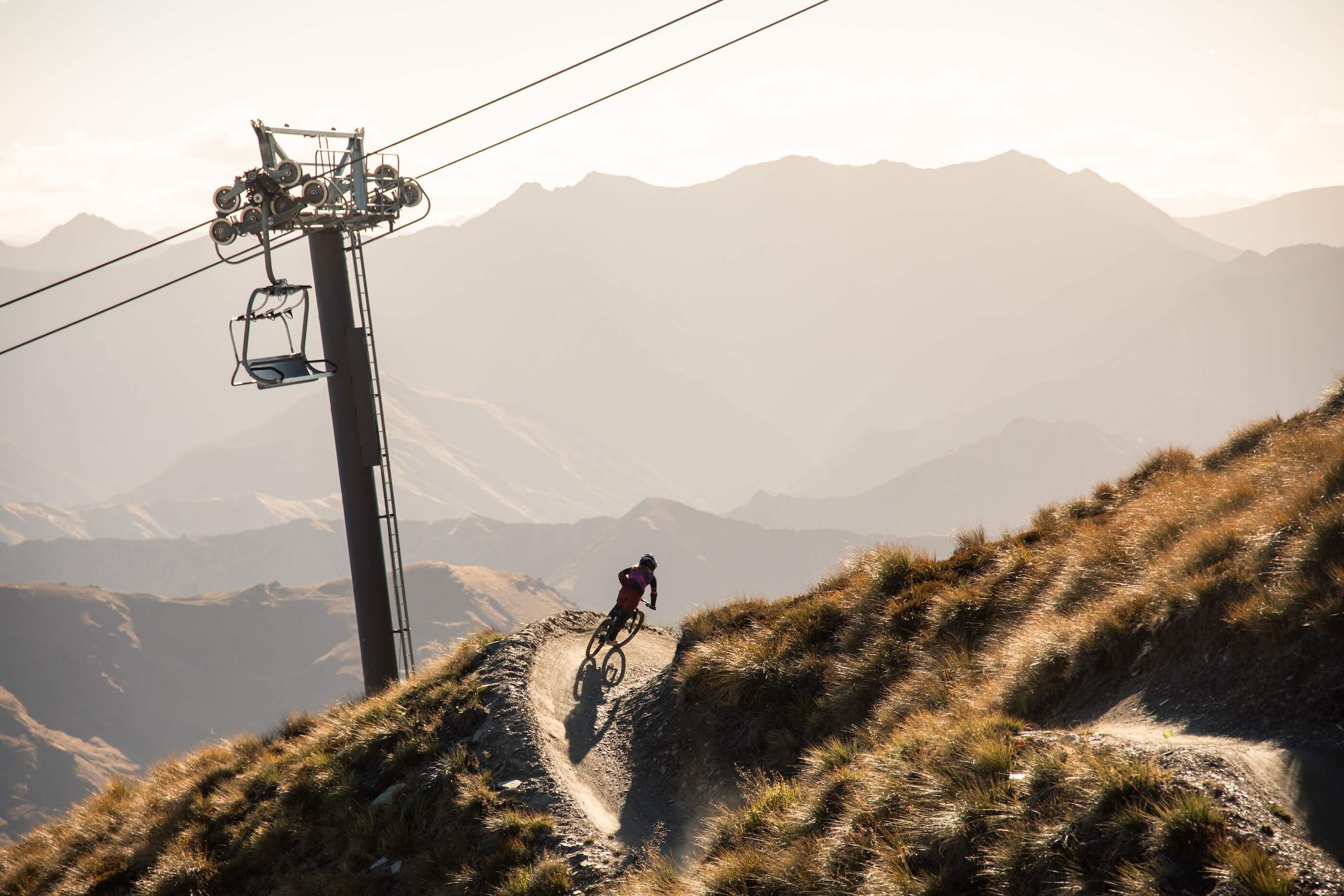Bicyclist rides mountain trail near ski lift with misty peaks in background at sunset.