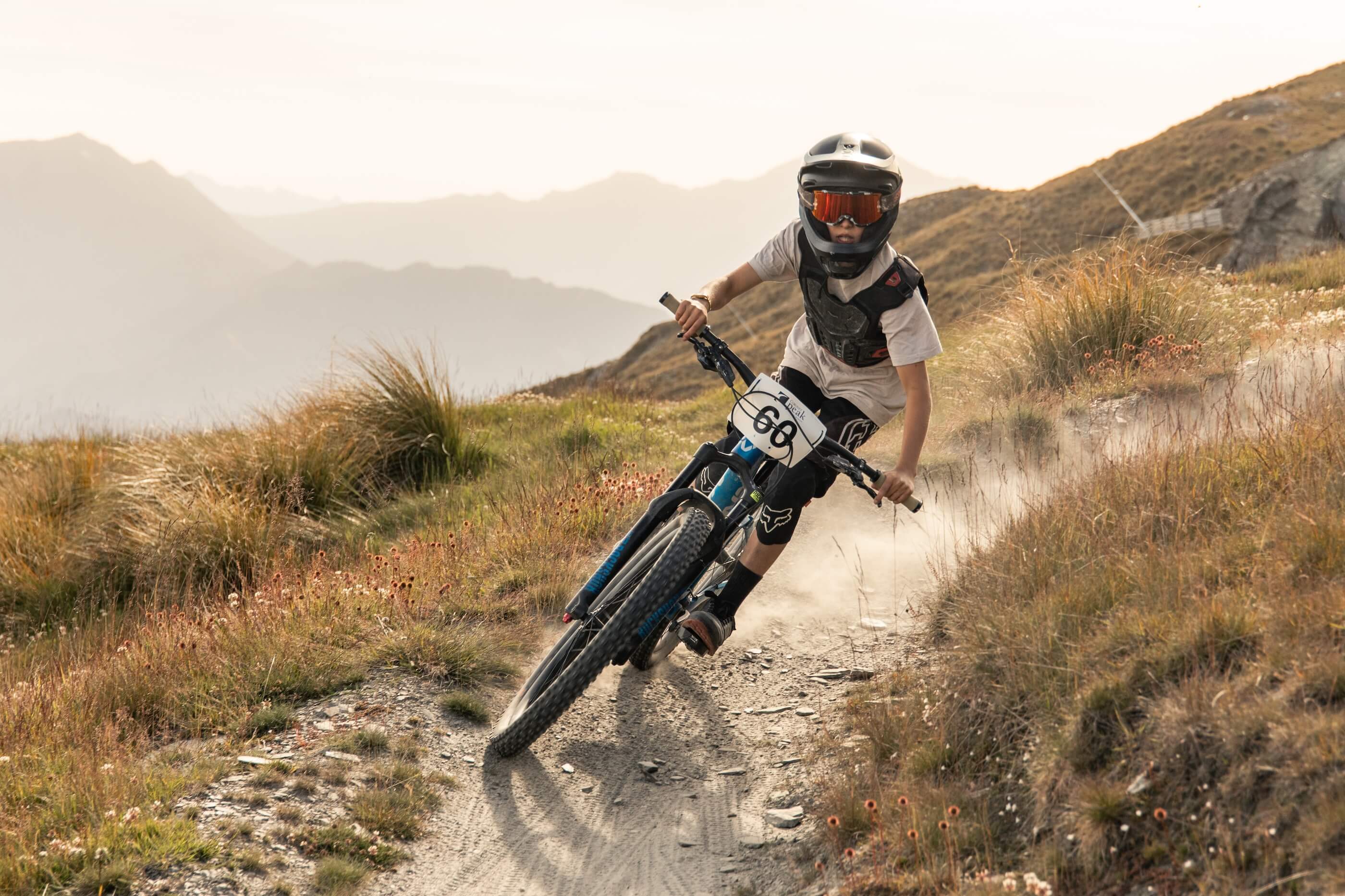 Cyclist wearing helmet and protective gear racing on a dusty mountain trail at sunset.