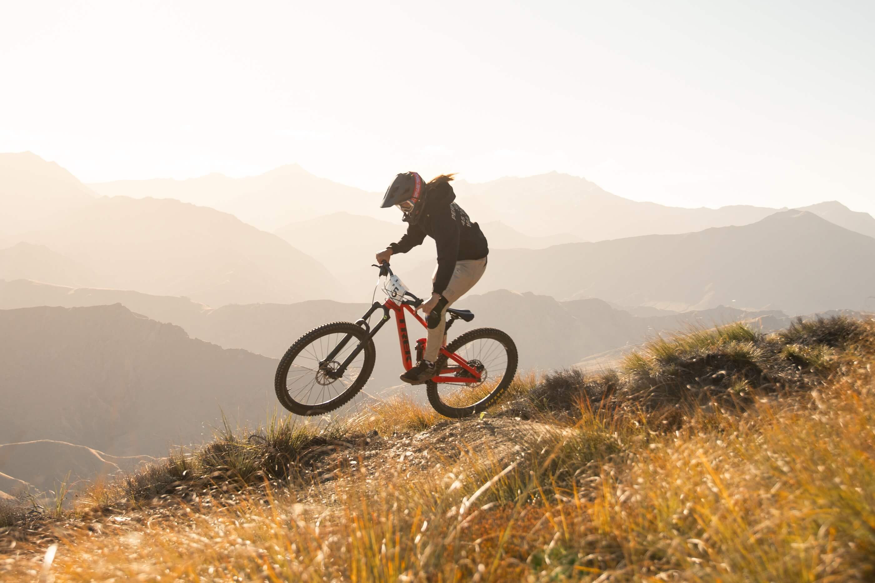 Mountain biker wearing a helmet rides uphill on a trail with mountains and grass at sunrise.