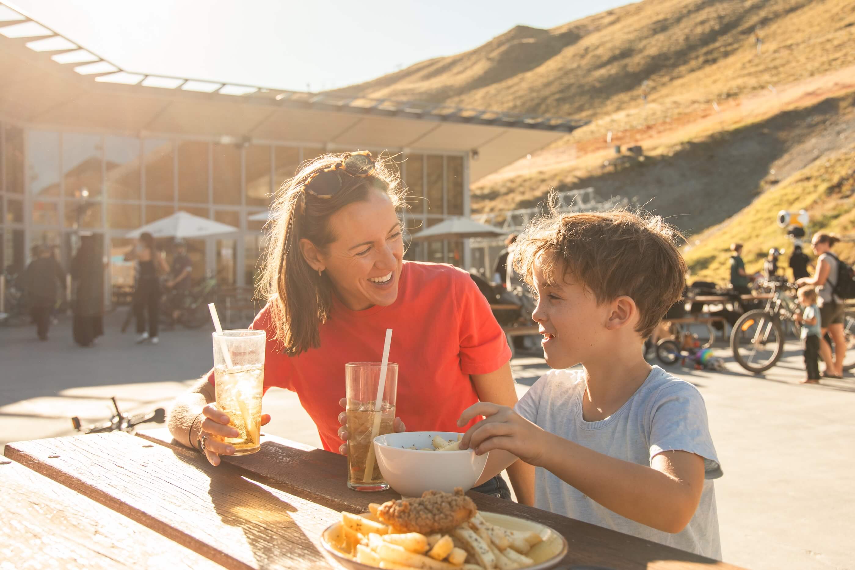 Smiling woman and boy sitting outdoors at a table with drinks and food in a sunny setting.