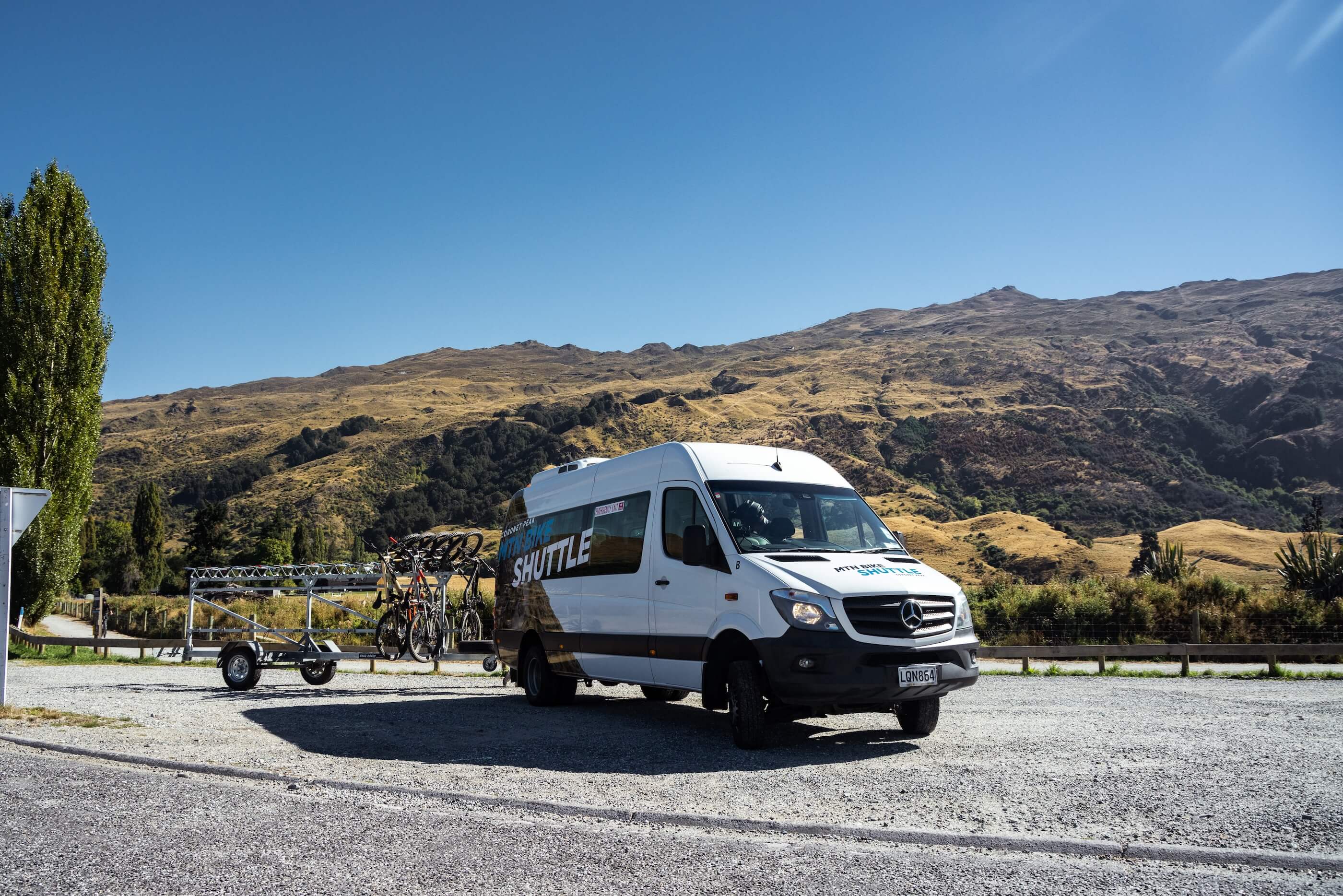 White shuttle van with mountain bike racks carrying bikes in a gravel lot near hills on a sunny day.