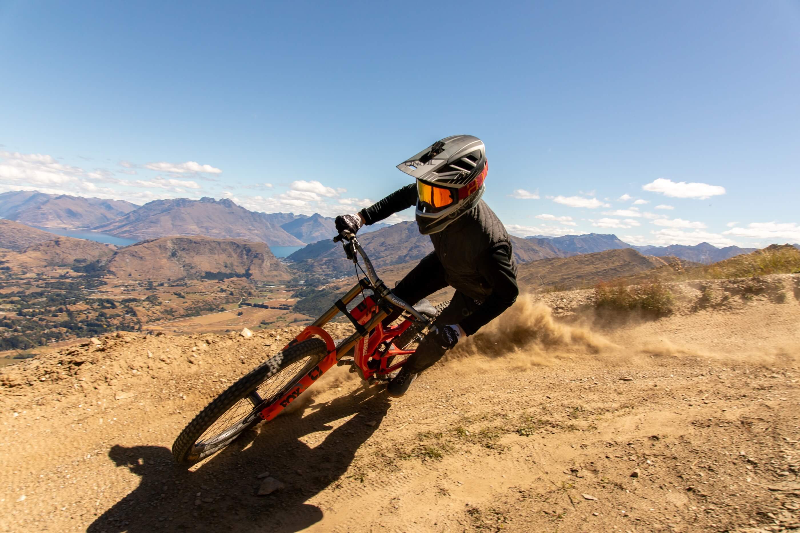 Mountain biker in black gear and helmet sharply turning on dusty trail with mountains in background.