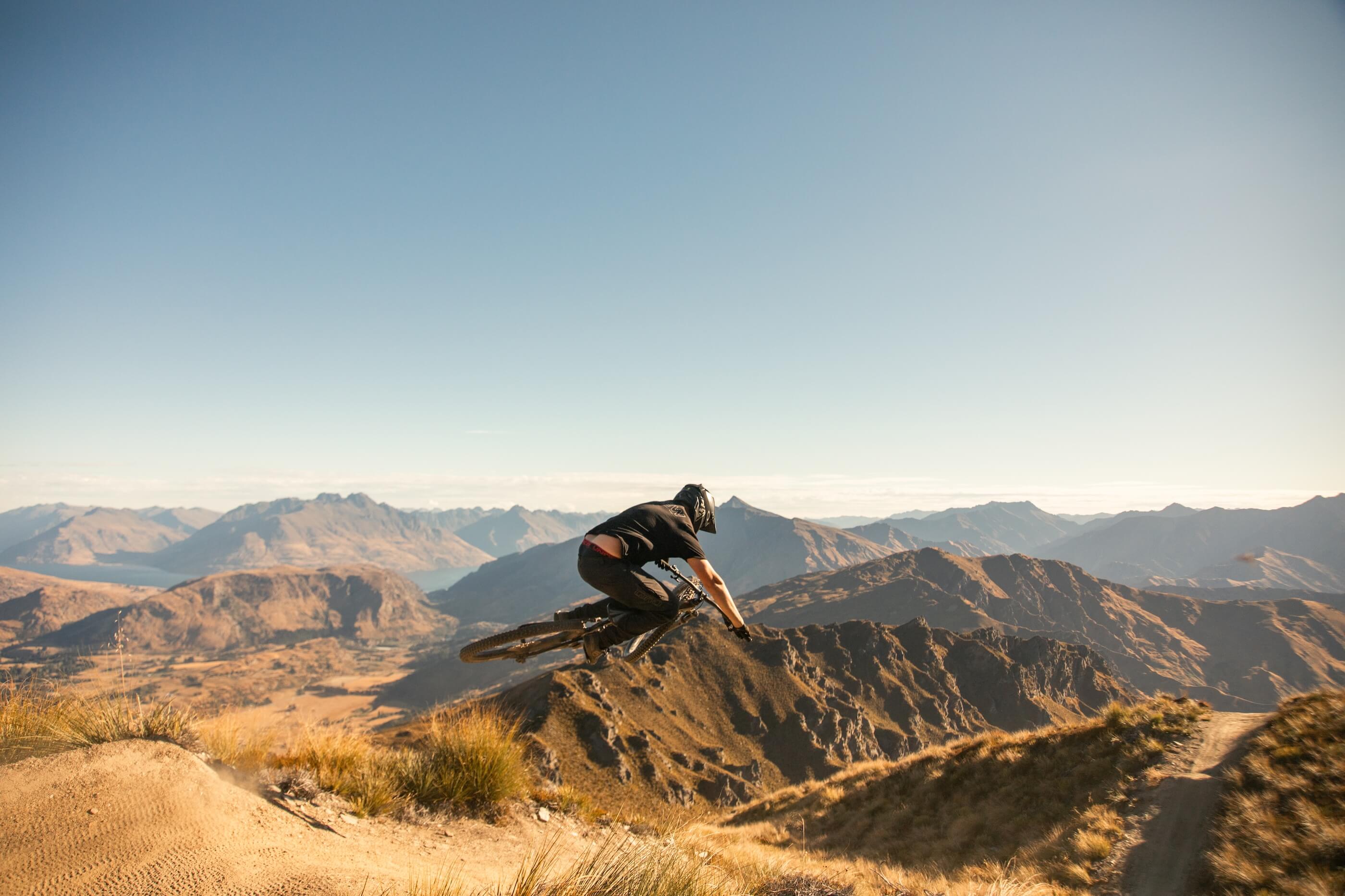 Mountain biker in mid-air over a dirt trail with rocky mountains under clear sky.