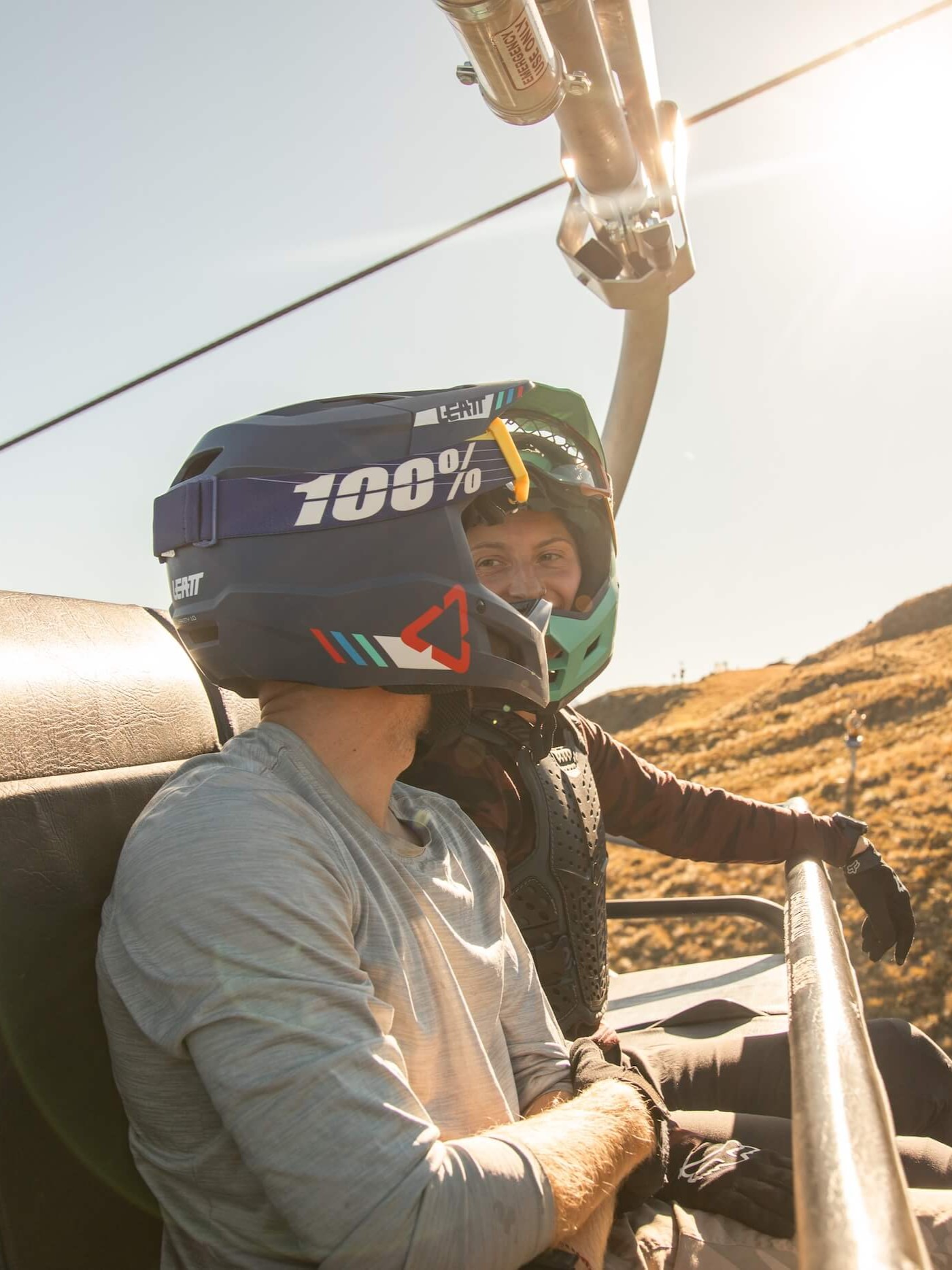 Two people in full-face helmets sitting on a ski lift with sunlit hills in the background.