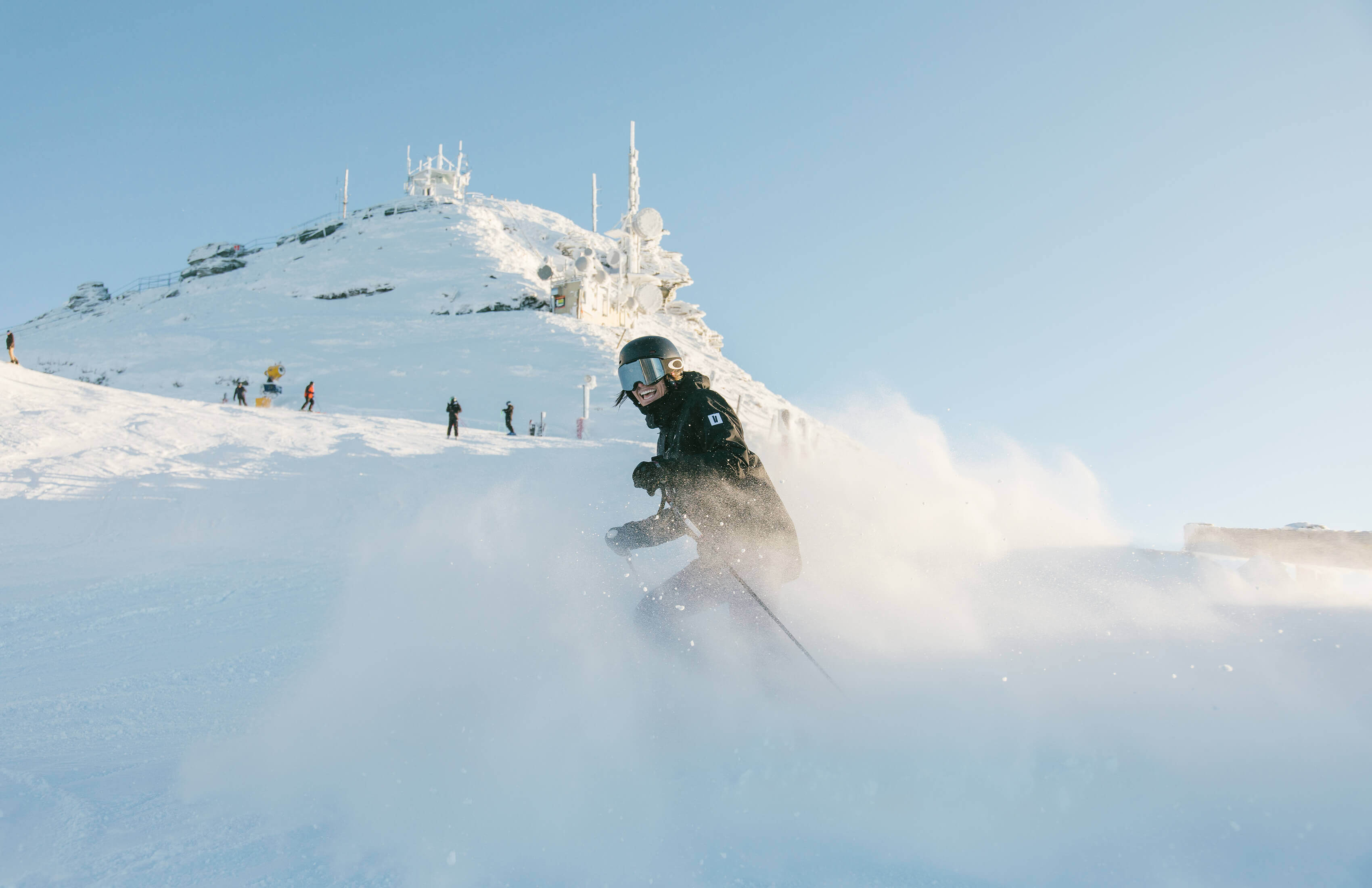 Smiling skier in black gear skiing down a snowy slope with mountain and clear sky in background.