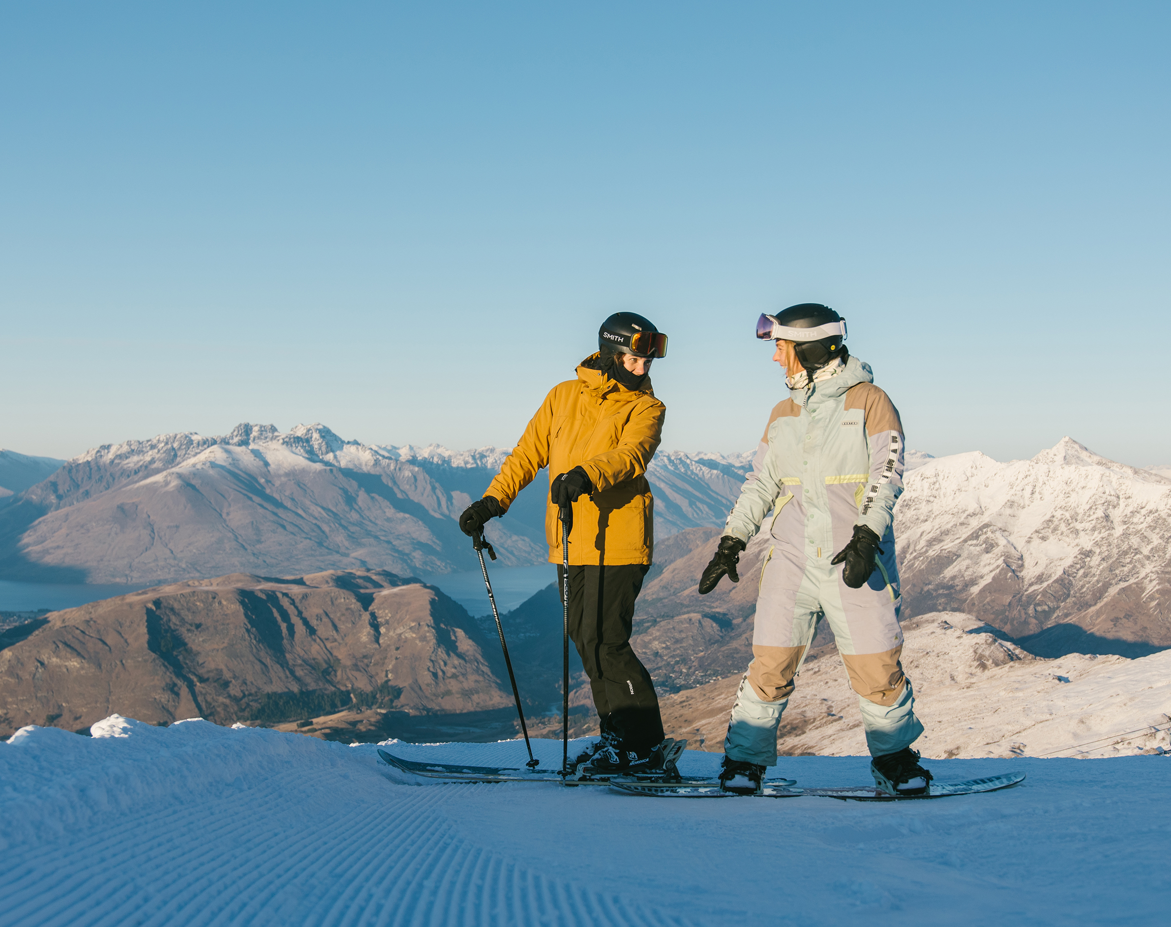 Two skiers chatting on a snow slope with snow-covered mountains and clear blue sky behind them.