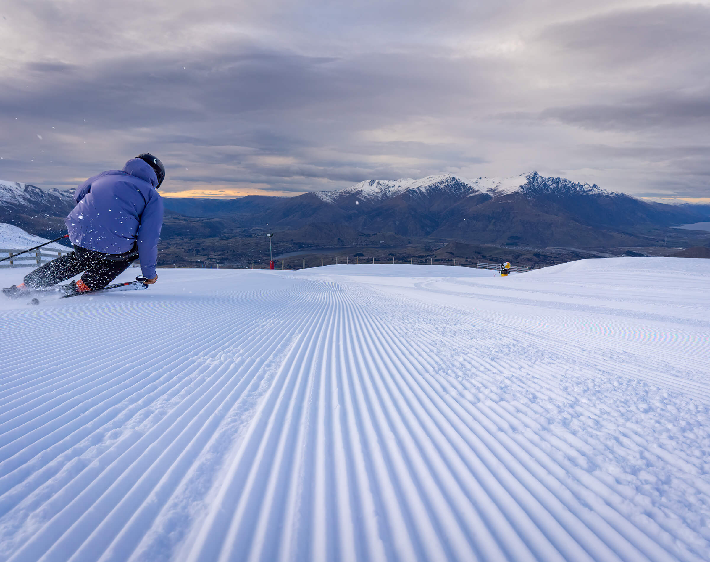 Skier in purple jacket carving down groomed slope with snowy mountains under cloudy sky.