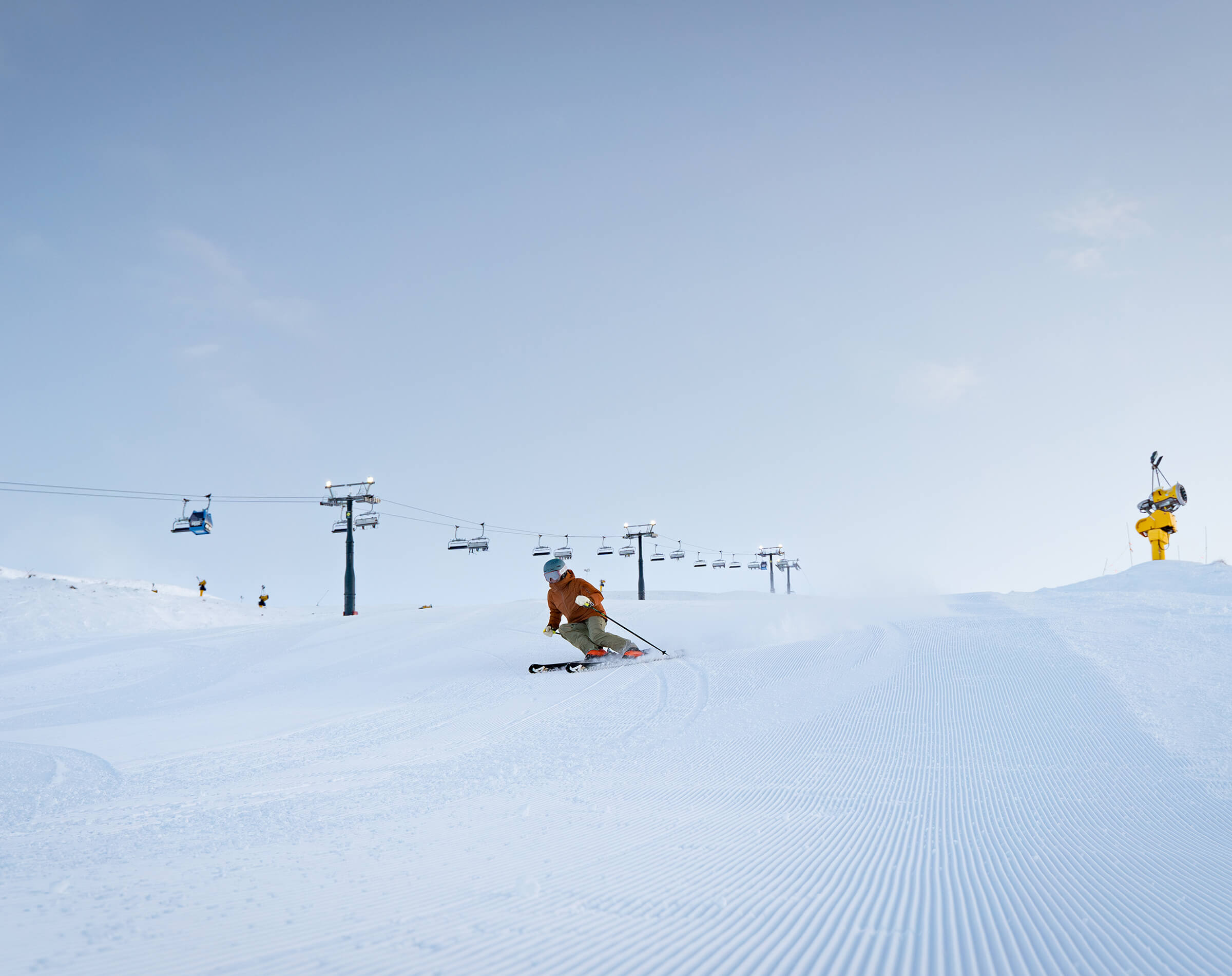 Skier in orange jacket skiing on groomed snow slope with ski lift and clear sky in background.