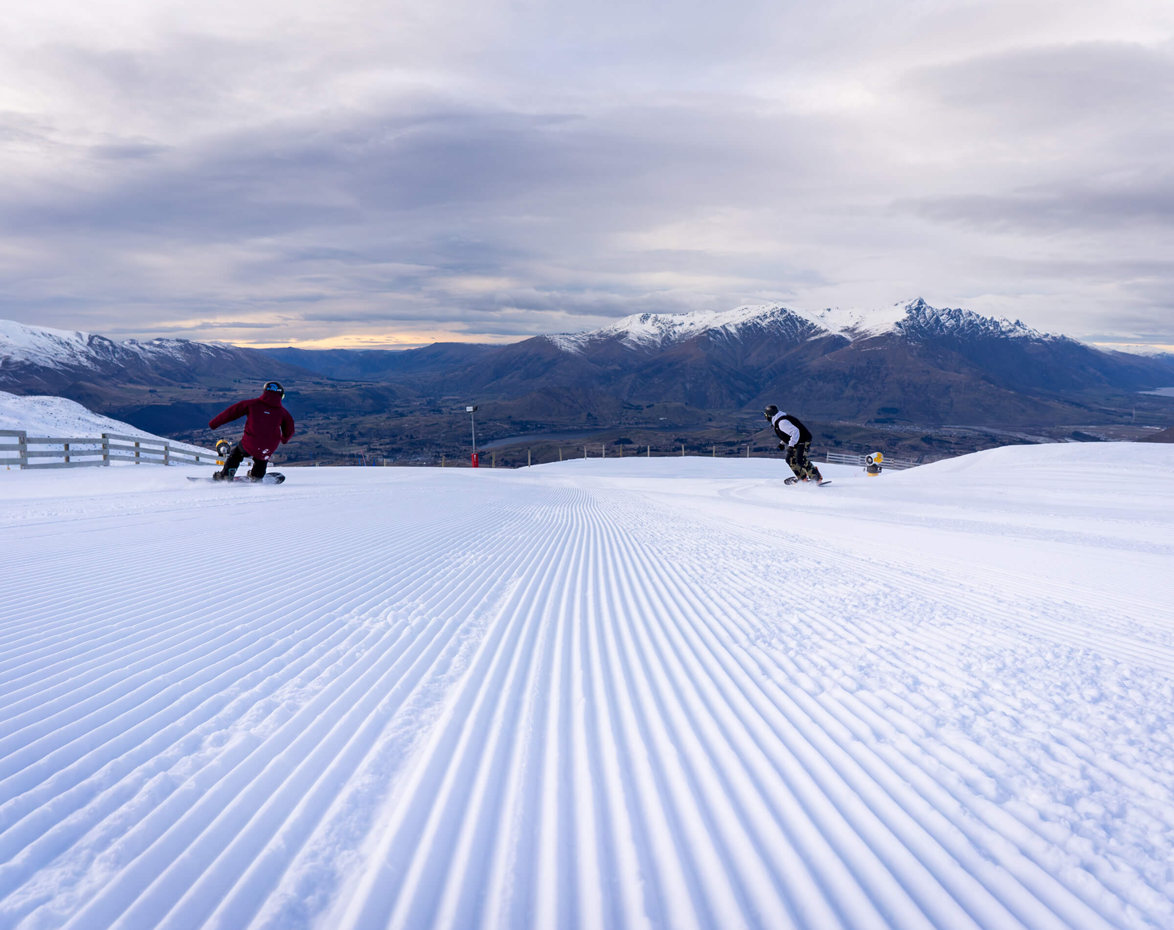 Two snowboarders riding down freshly groomed snowy slope with mountain views under cloudy sky.