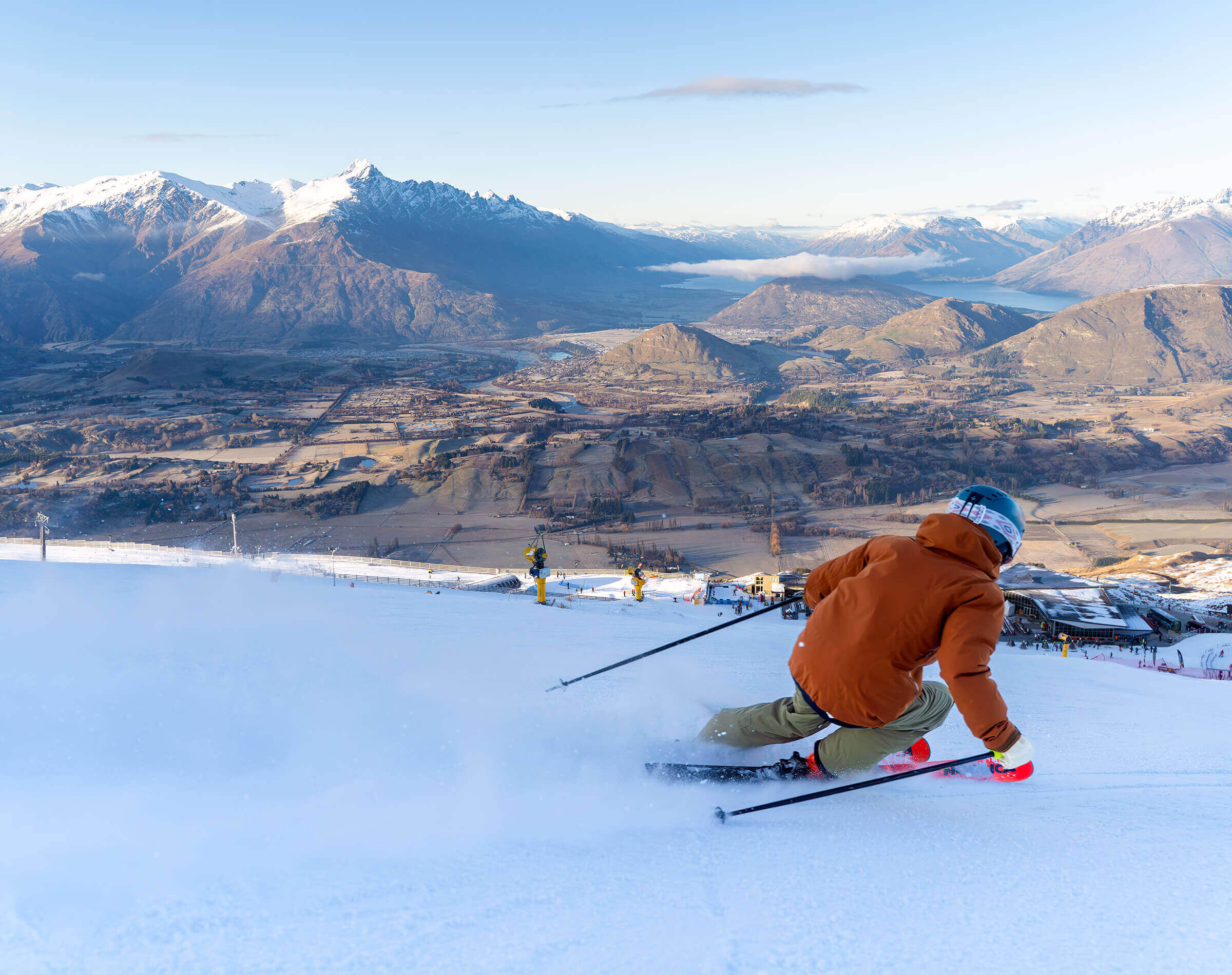 Skier in orange jacket carving down snowy slope with mountains in the background.