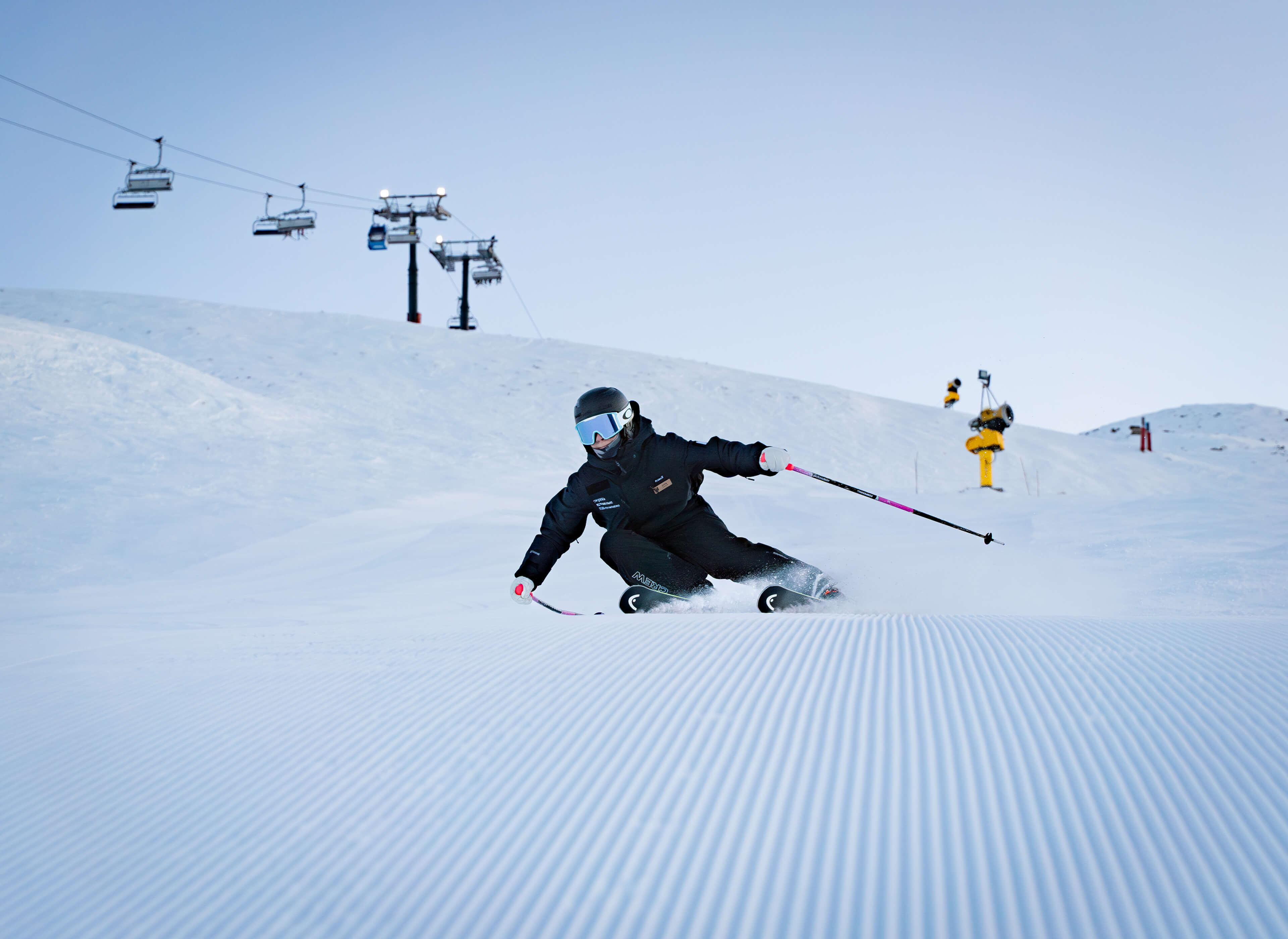 Skier in black gear carving a turn on groomed snowy ski slope under clear sky.